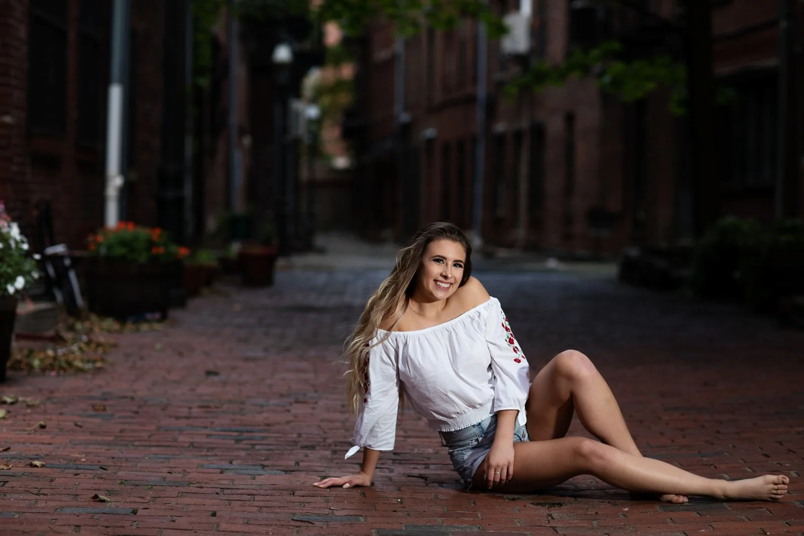 Young woman sitting on a brick alleyway, smiling, wearing an off-the-shoulder white blouse with floral embroidery and denim shorts, with potted plants and brick buildings in the background during dusk.