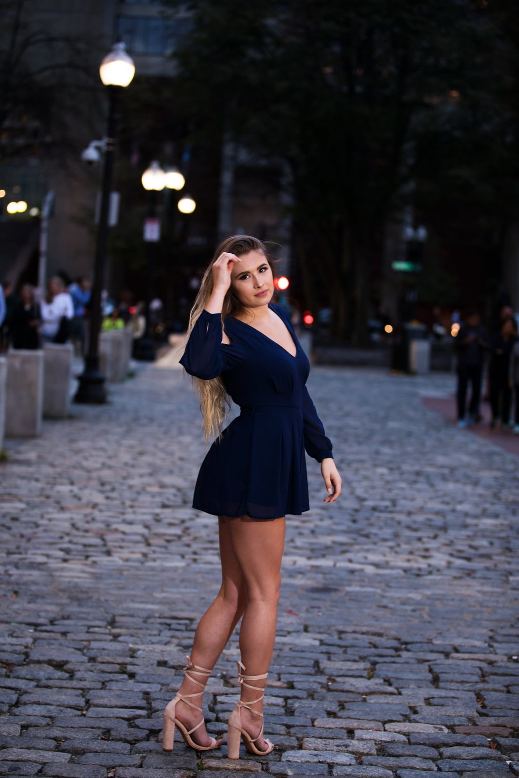 A woman standing on a cobblestone street at dusk, wearing a navy blue dress and beige high-heeled sandals, with her left hand touching her hair and looking at the camera.