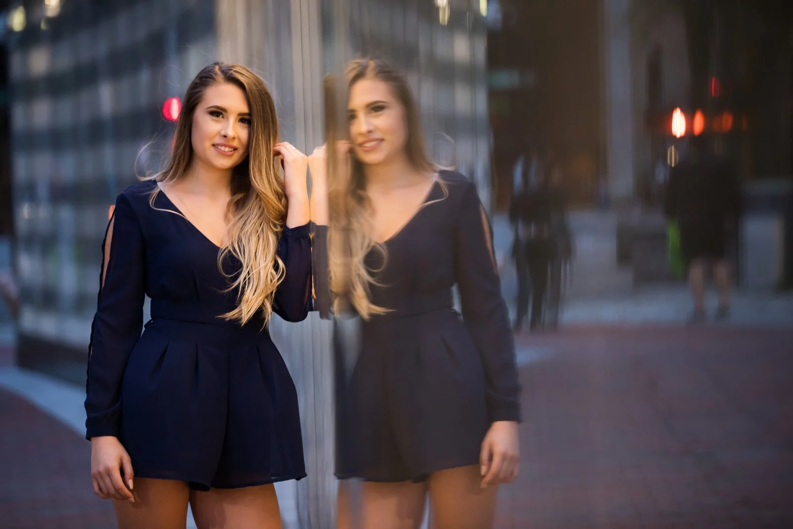 A young woman with long blonde hair in a navy blue dress standing next to a reflective glass wall, with her reflection visible in the glass