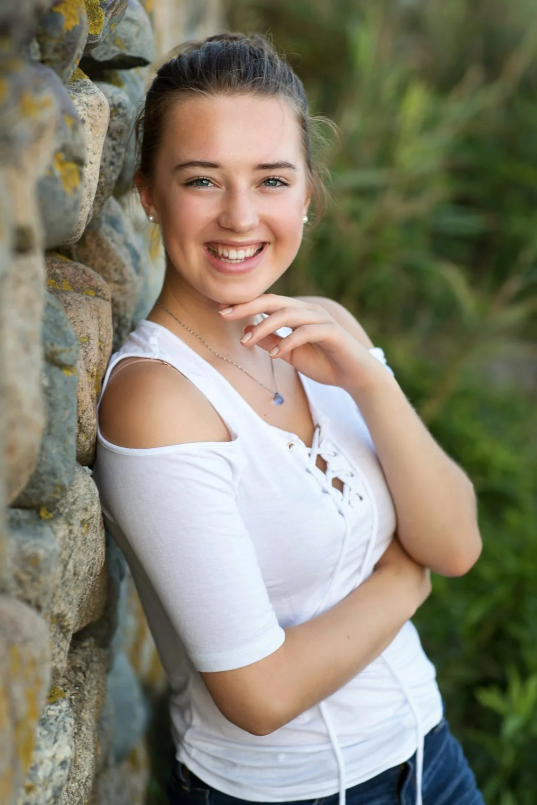 A young woman smiling and leaning against a stone wall outdoors, wearing a white cut-out shoulder top and jeans.