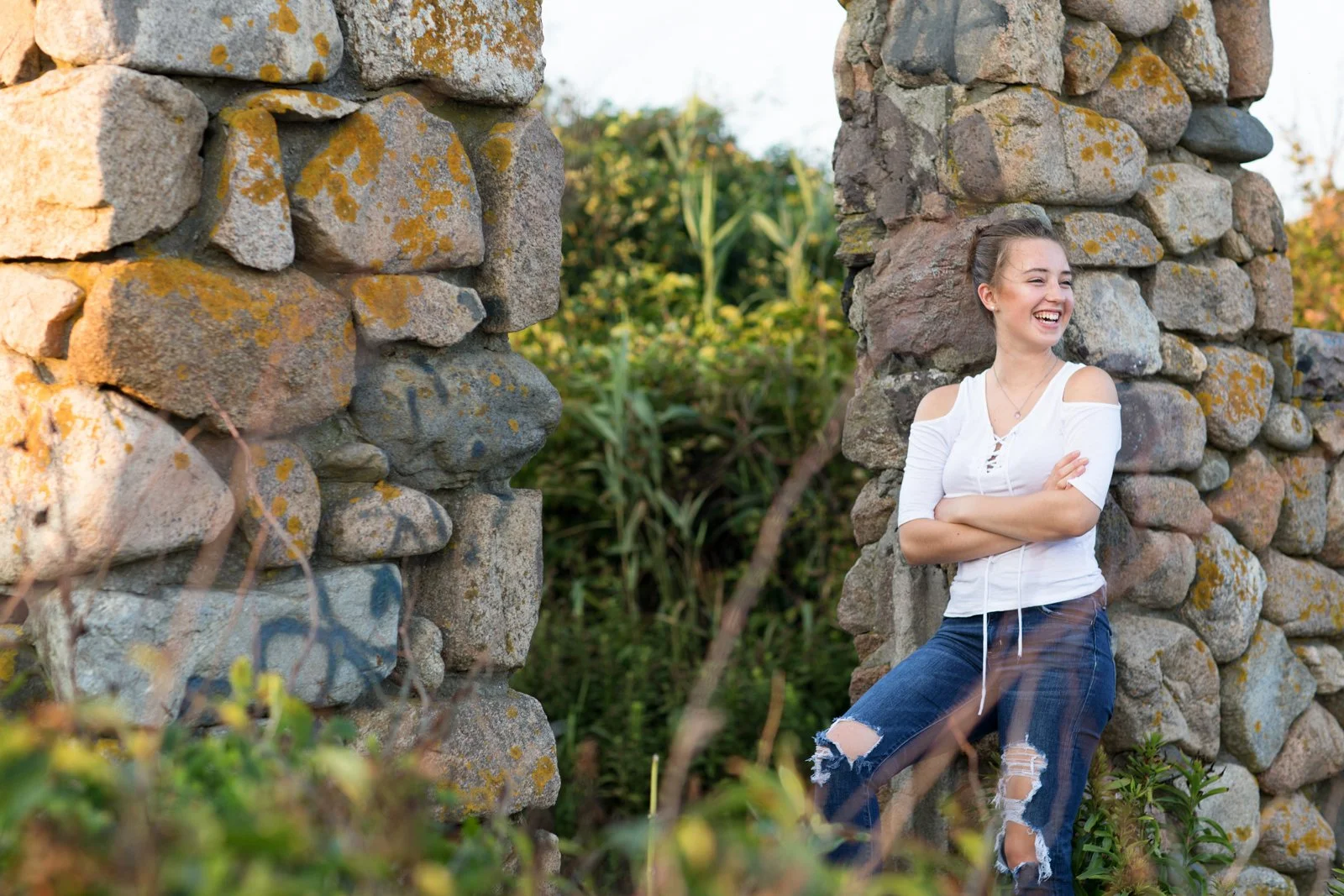 A young woman with a white top and ripped jeans, smiling and standing outdoors between two large stone structures, surrounded by greenery.