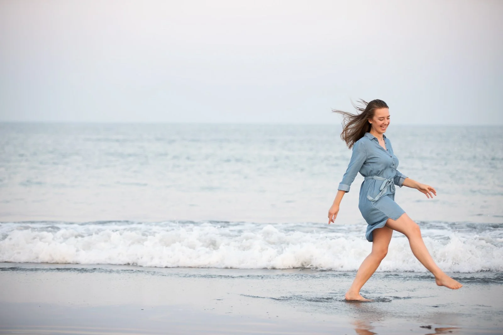 A woman wearing a denim dress walking barefoot on the beach near the shoreline with the ocean in the background, smiling and enjoying the moment.