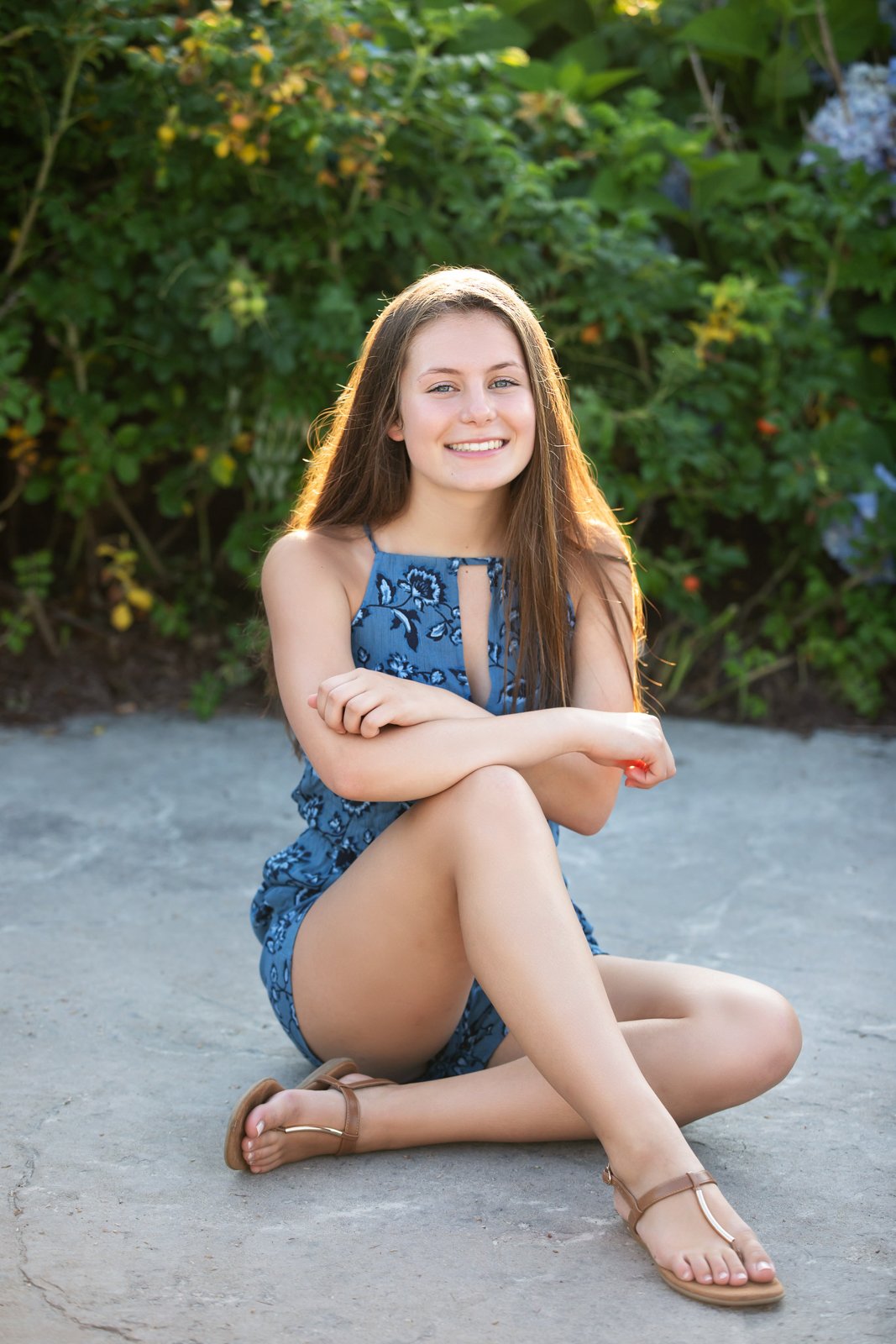 A young woman with long brown hair, wearing a blue floral sleeveless dress, sitting cross-legged on a concrete surface outdoors with leafy greenery and yellow and blue flowers in the background, smiling at the camera.
