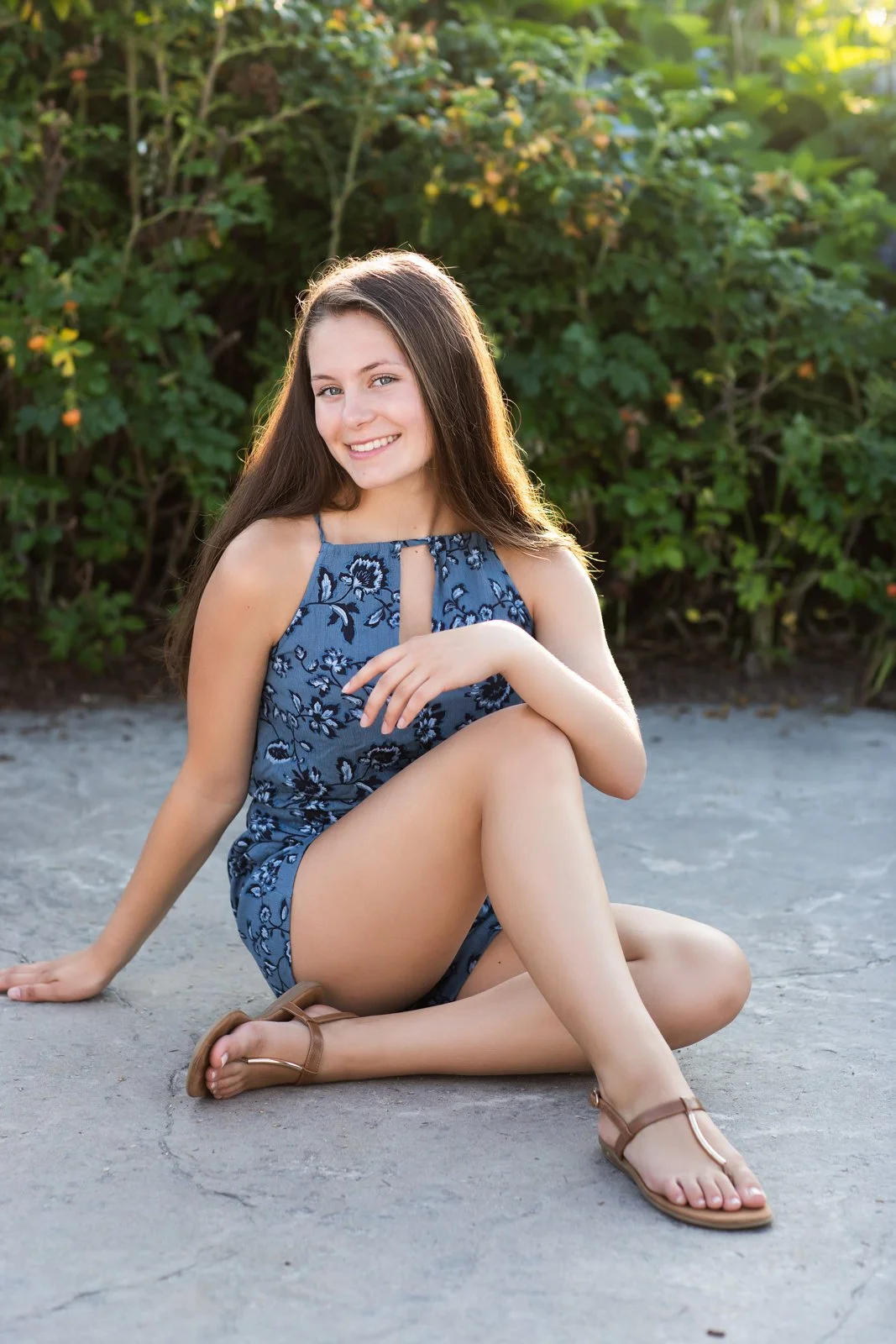 A young woman with long brown hair, smiling, sitting on the ground outside in front of green bushes, wearing a blue floral dress and brown sandals.