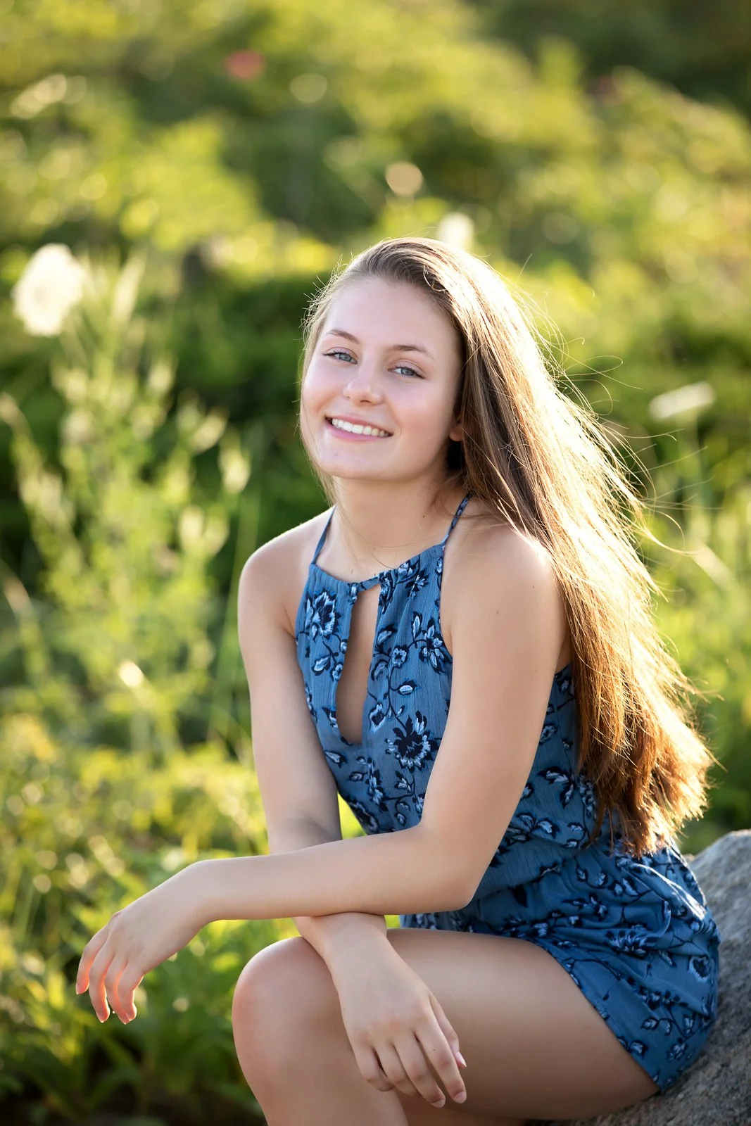 A young woman with long, light brown hair, wearing a blue floral sleeveless dress, sitting outdoors on a rock, smiling with sunlight illuminating her face and hair, with a green, blurred natural background.