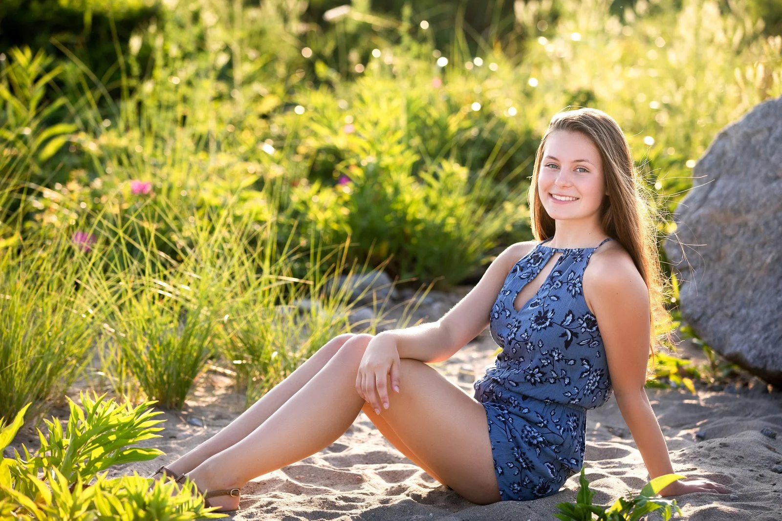 Young woman sitting on the sandy beach with greenery in the background during sunset, smiling, wearing a blue floral sleeveless dress.