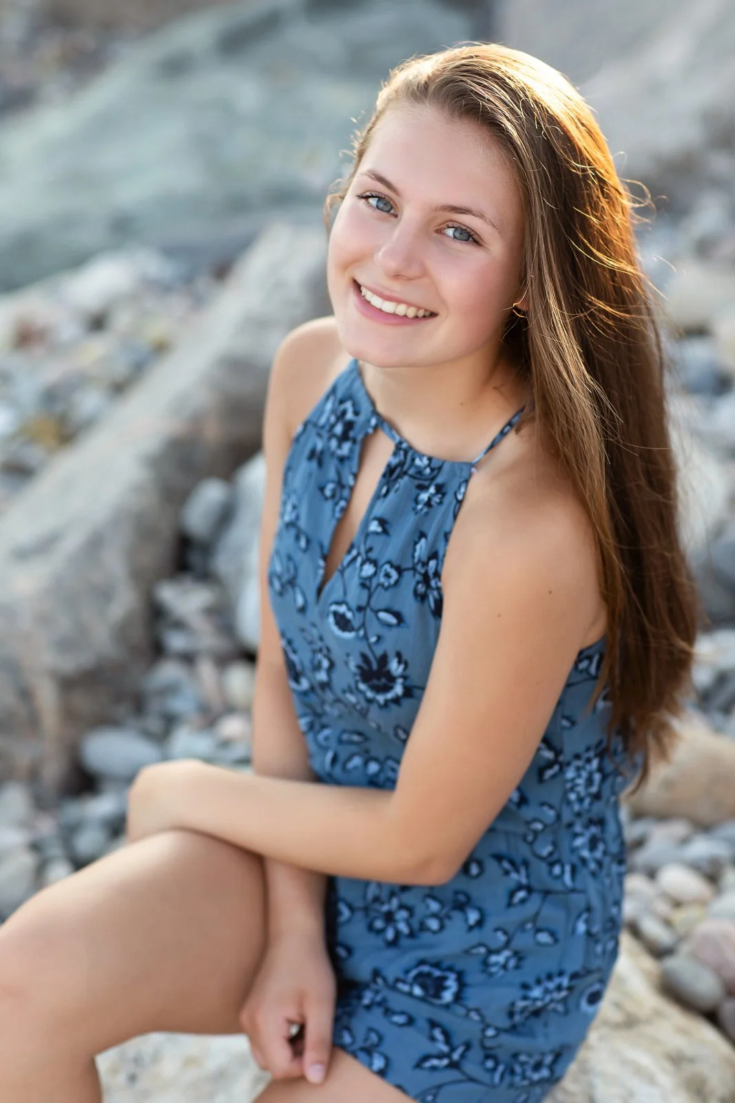 A young woman with long brown hair and blue eyes, smiling, wearing a blue floral sleeveless dress, sitting on rocks near water.
