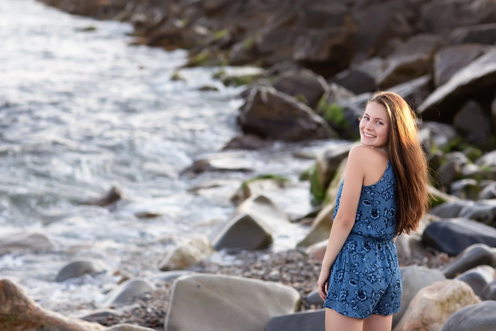 A young woman with long brown hair smiling at the camera, wearing a blue floral sleeveless romper, standing on a rocky beach near the water.