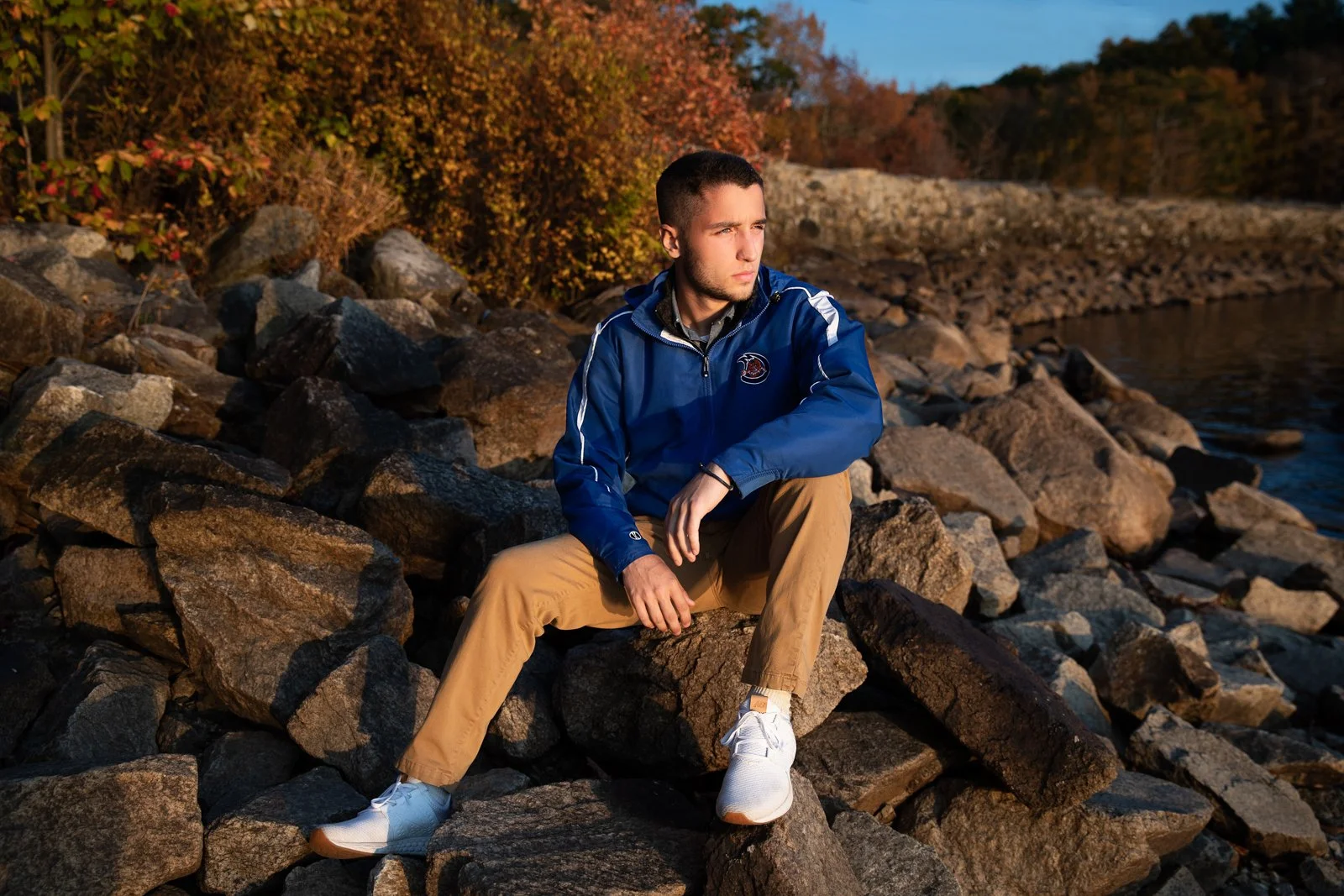 A young man sitting on rocks by a body of water with autumn foliage in the background, wearing a blue jacket, tan pants, and white sneakers, looking contemplatively to the side.