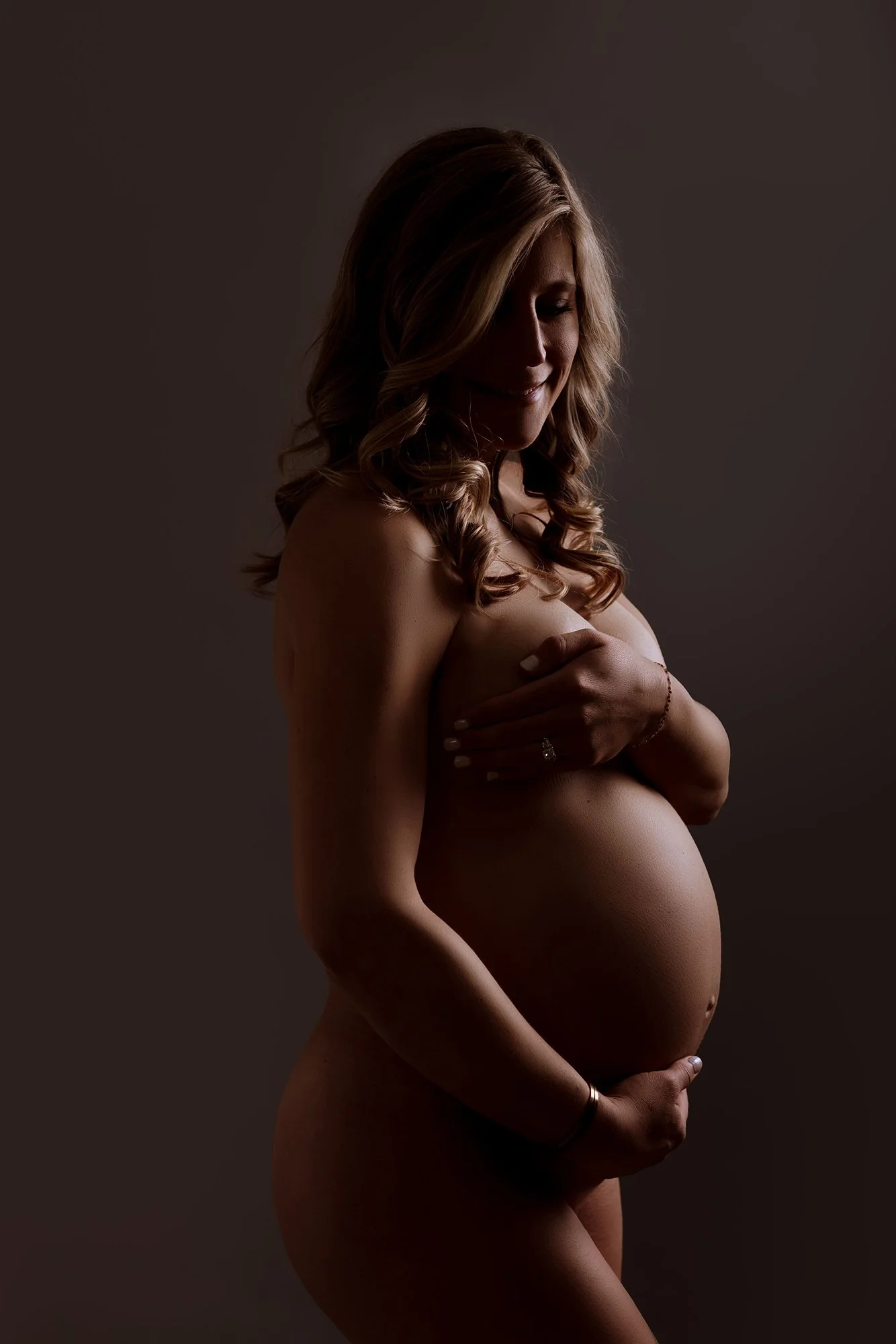 A pregnant woman with long, wavy hair, smiling softly, covering her breasts with one hand and supporting her belly with the other, against a dark background.