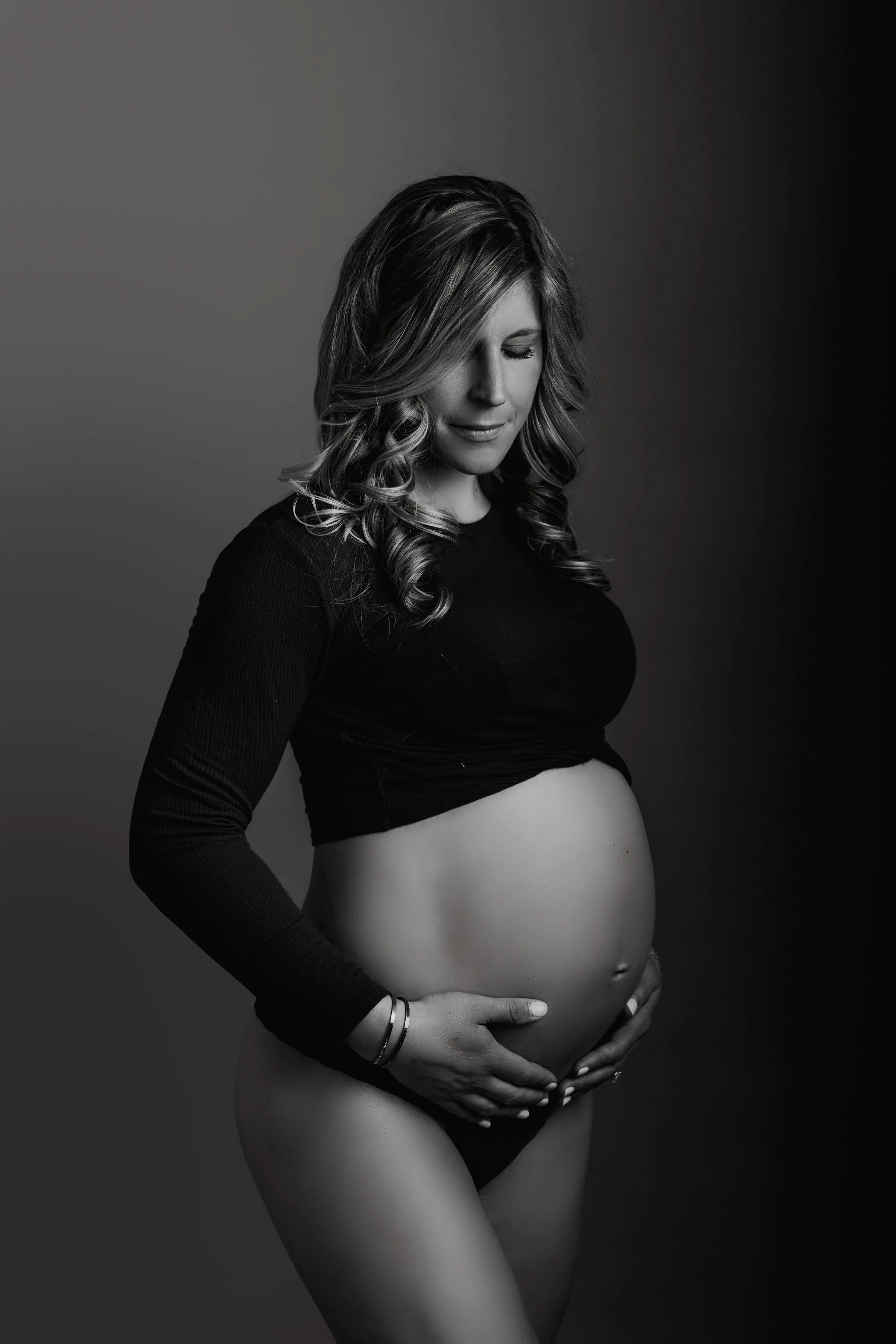 A pregnant woman with wavy hair, wearing a black long-sleeved top and underwear, holds her belly and looks down, set against a plain background.