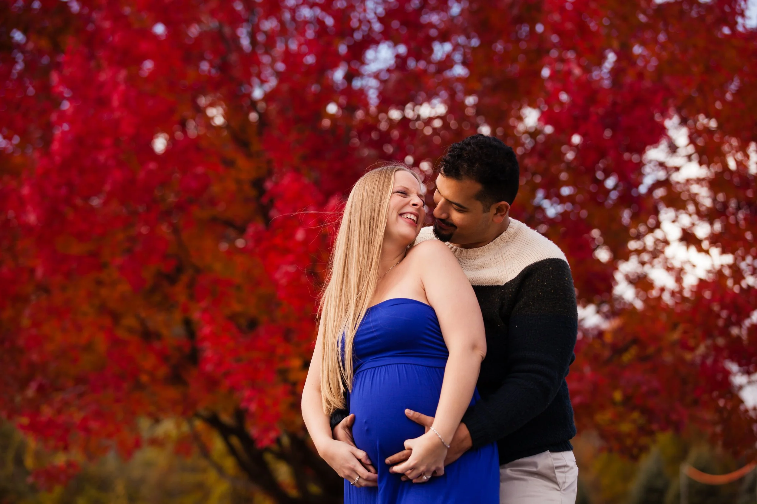 A smiling pregnant woman in a blue dress and a man are holding her belly and smiling at each other outdoors in front of a background of bright red autumn leaves.