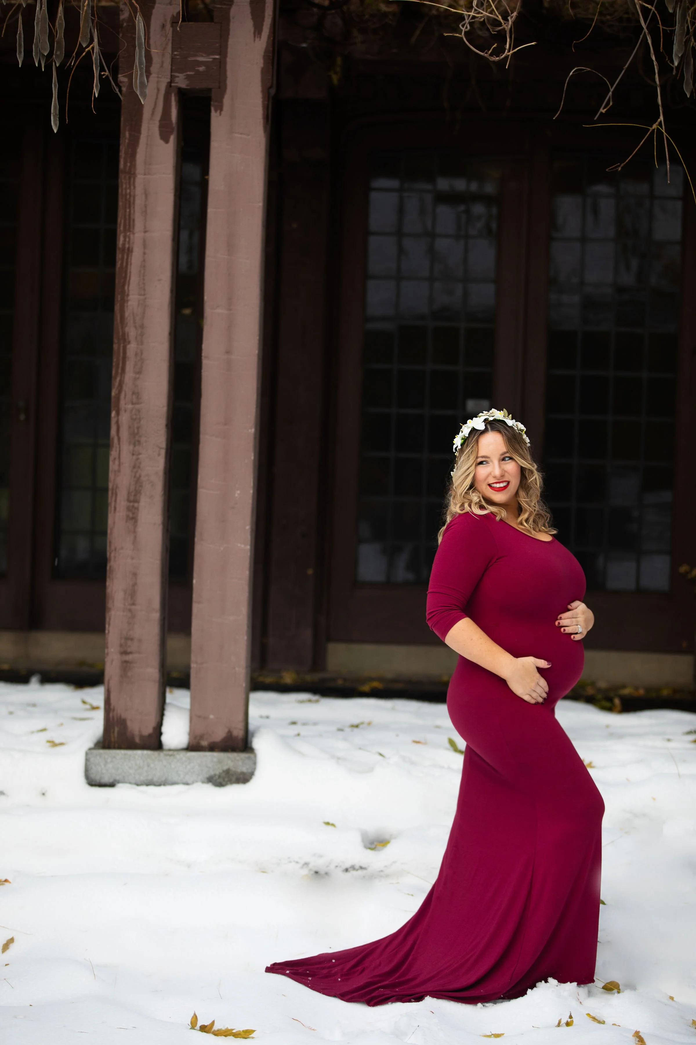 A pregnant woman in a long, maroon dress and floral crown stands outdoors on snow, smiling and holding her belly, in front of a wooden background.