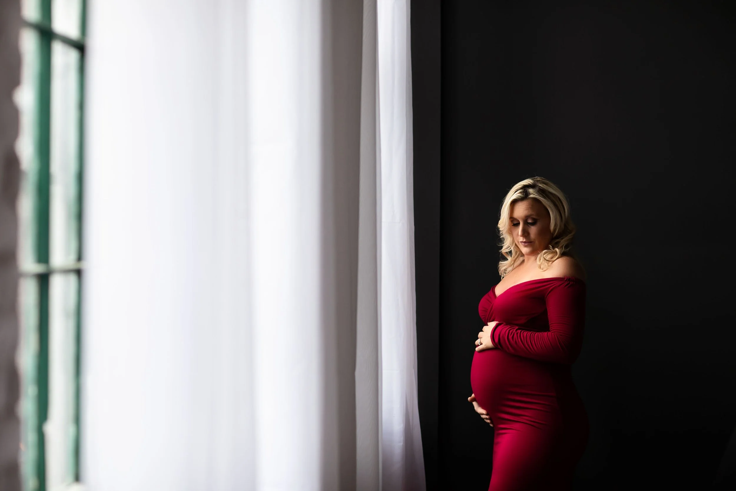 Pregnant woman in a red dress standing by a window with white curtains, looking down, dark background.