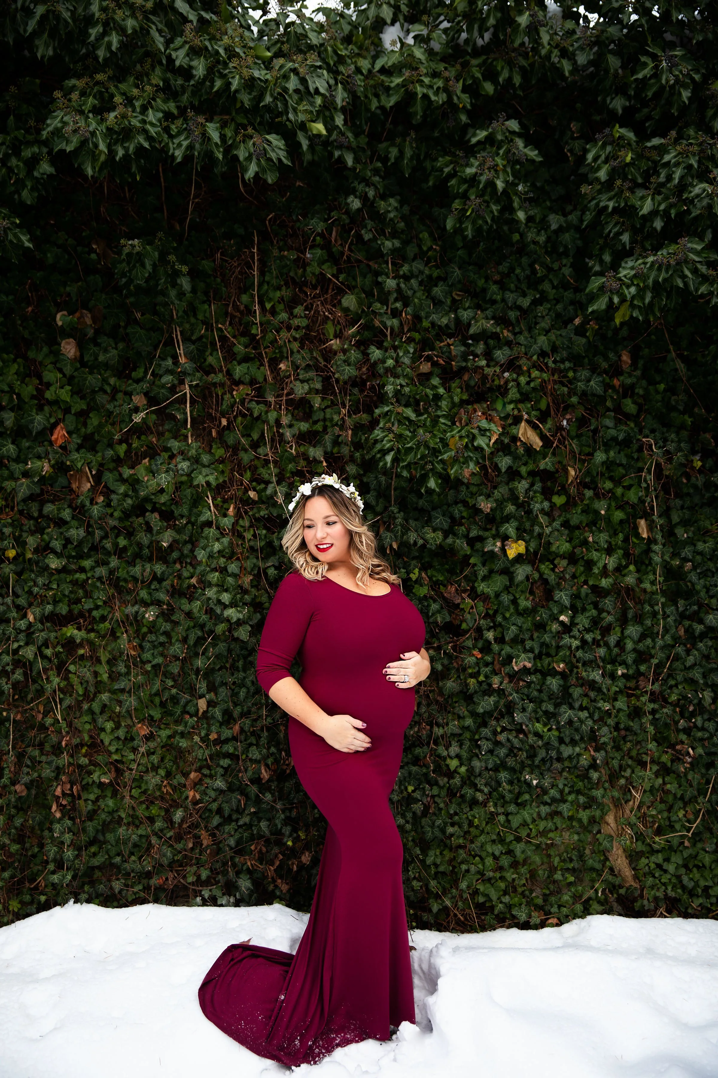A pregnant woman in a burgundy gown standing on snow in front of a dense green ivy-covered wall, wearing a floral crown, looking down and smiling.