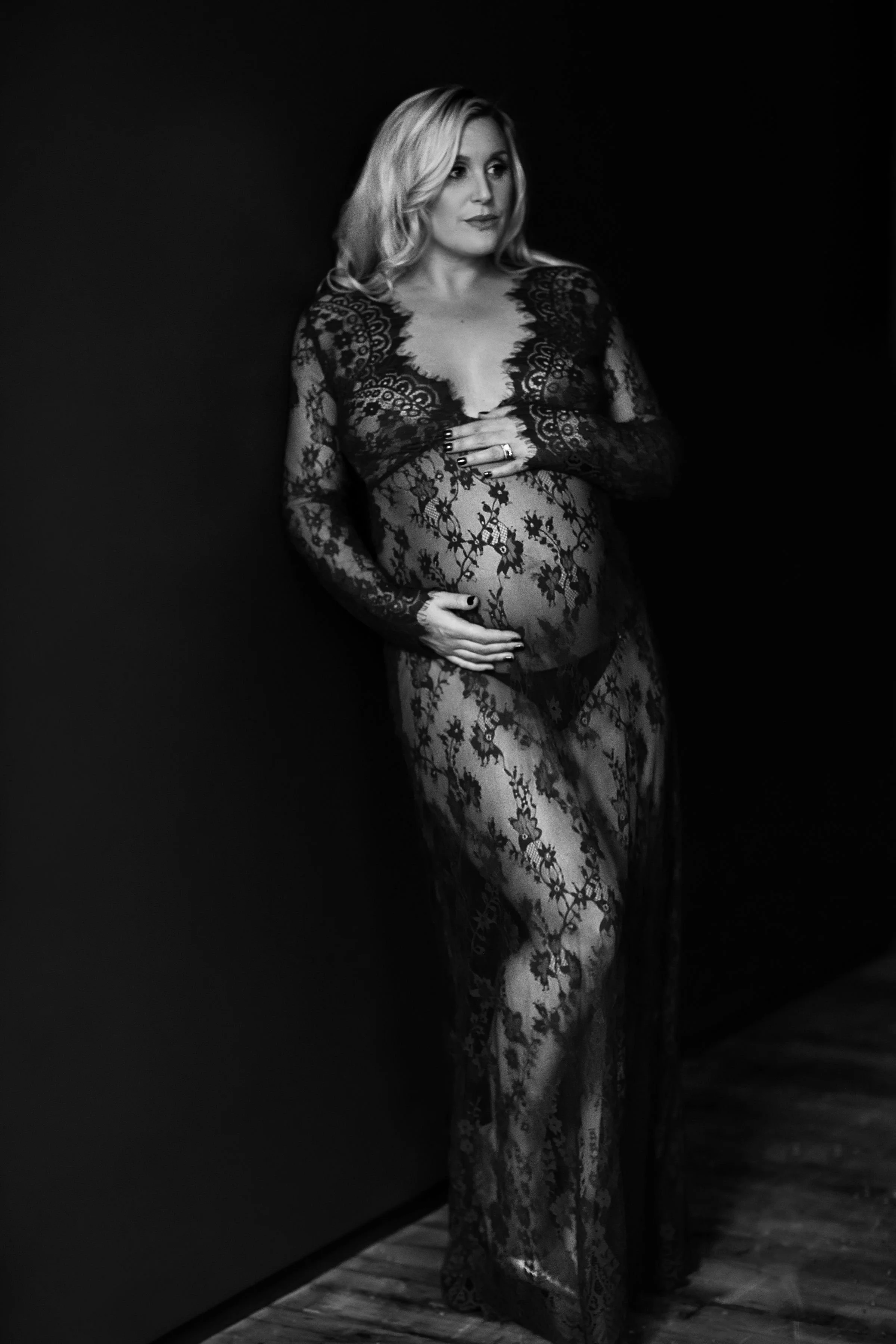 A pregnant woman dressed in a black lace gown, standing against a dark background, with one hand on her belly and the other resting on her chest.