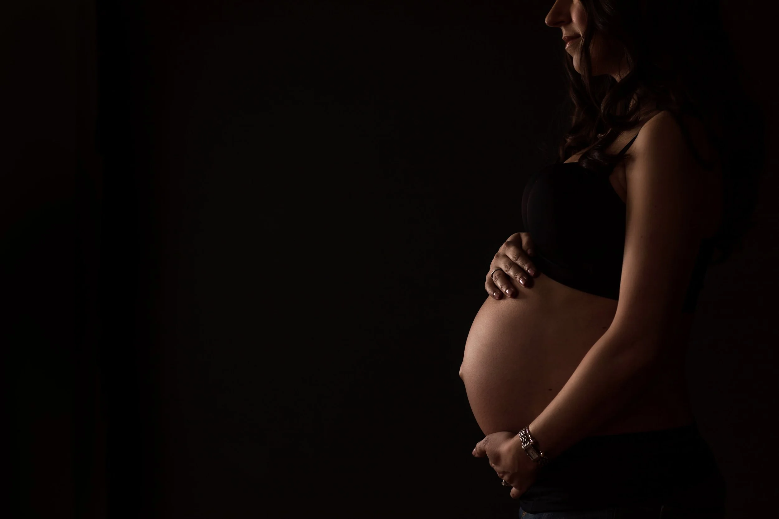 Profile of a pregnant woman in a dark setting, with her face partially visible, wearing a black top, with her hand on her stomach.