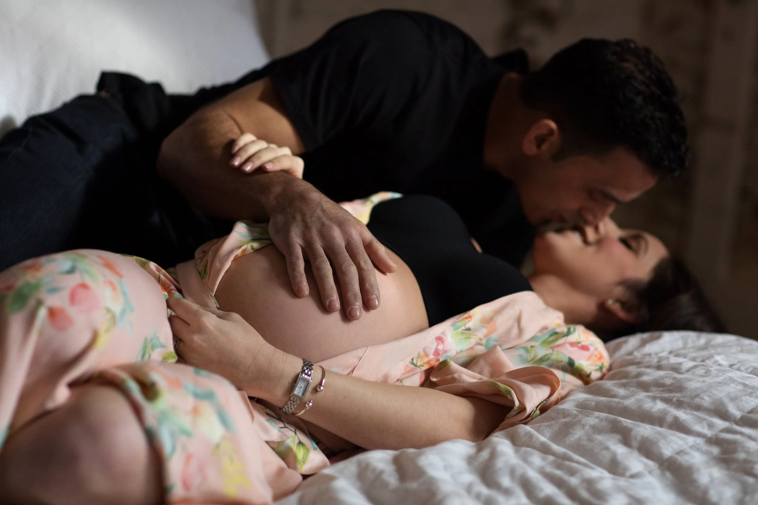 A couple sharing an intimate moment on a bed, with the woman pregnant, in a dimly lit room.