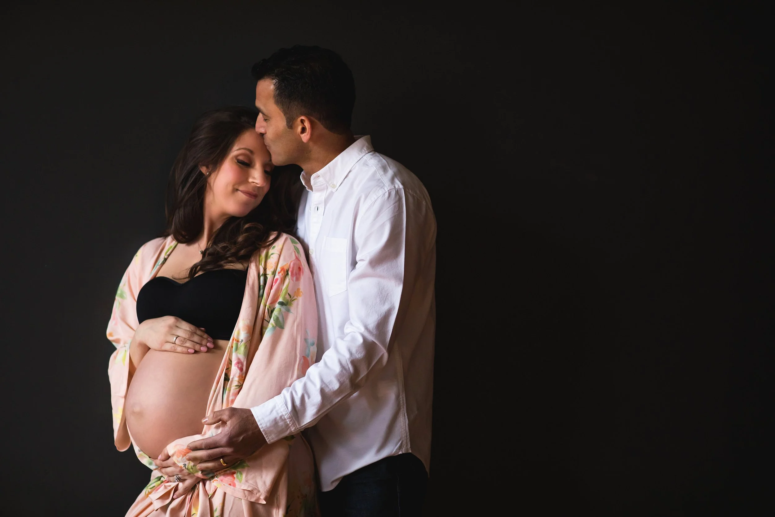 Pregnant woman with long dark hair, wearing a black top and floral robe, gently holding her belly, with a man in a white shirt embracing her and kissing her forehead against a black background.