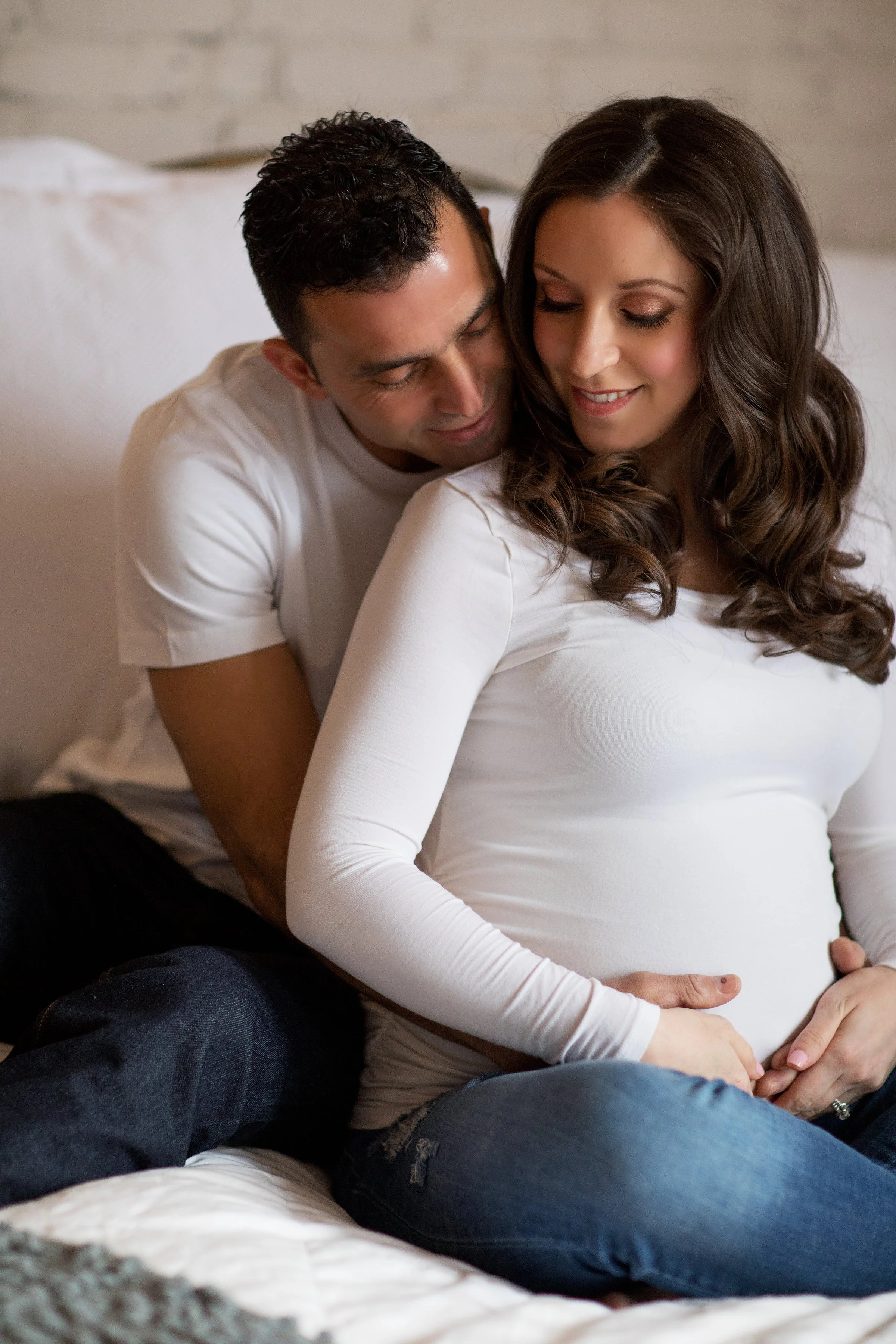 A pregnant woman sitting on a bed with a man behind her, both smiling and embracing each other, in a cozy bedroom setting.