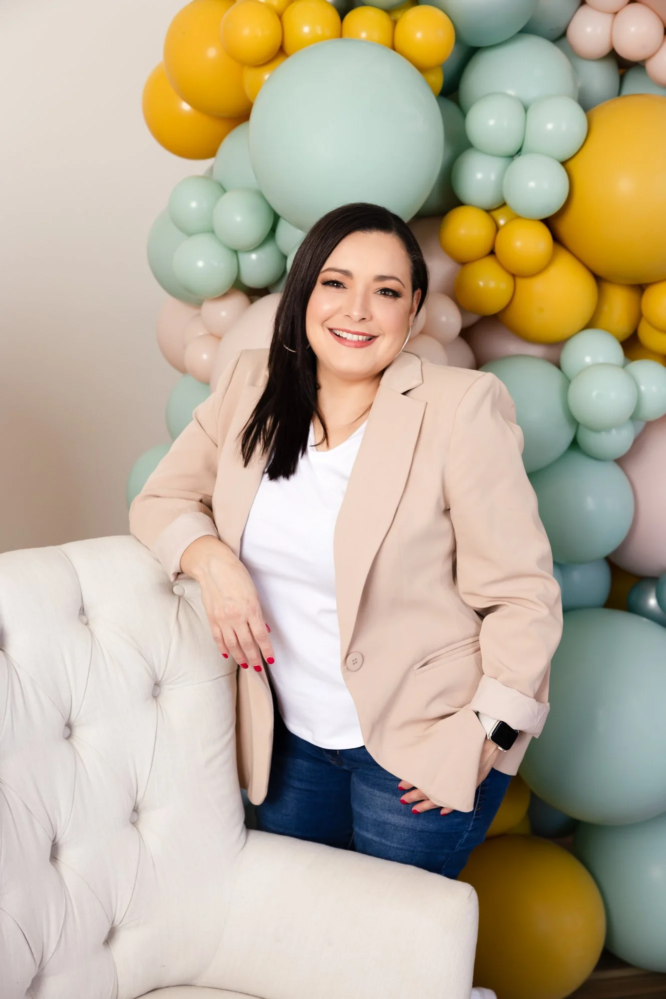 A smiling woman with black hair, wearing a beige blazer and white shirt, posing beside a cream-colored tufted chair, with a colorful balloon backdrop of yellow, mint, white, and soft pink balloons.