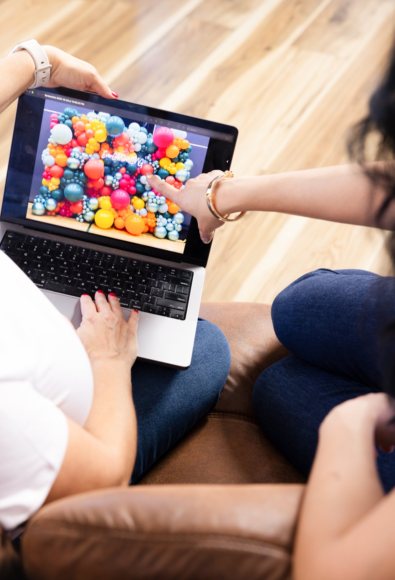 Two women sitting on a couch, one holding a laptop that displays a colorful abstract balloon artwork, and the other pointing at the screen.