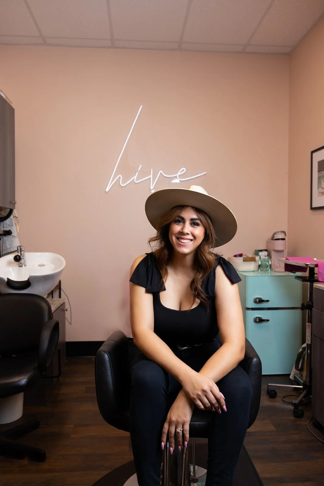 A woman sitting on a black chair in a salon, smiling, wearing a wide-brimmed hat and a black top. Behind her is a wall with a neon sign that says 'hype'. To her left is a sink and to her right is a small mint-colored refrigerator and a pink coffee ma