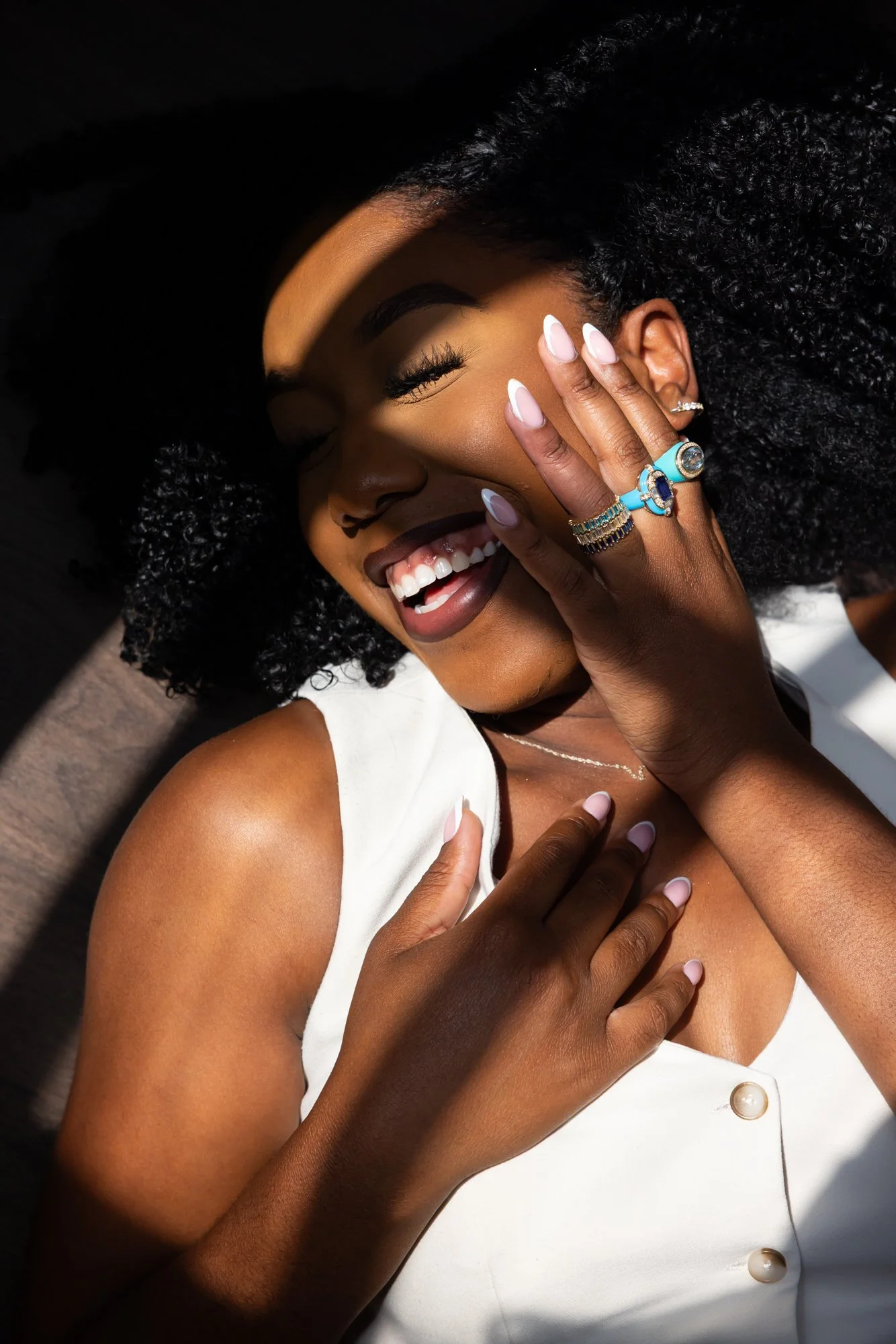 A smiling woman with curly hair and white top, covering her face with one hand, showing rings and jewelry, with sunlight casting shadows on her face.