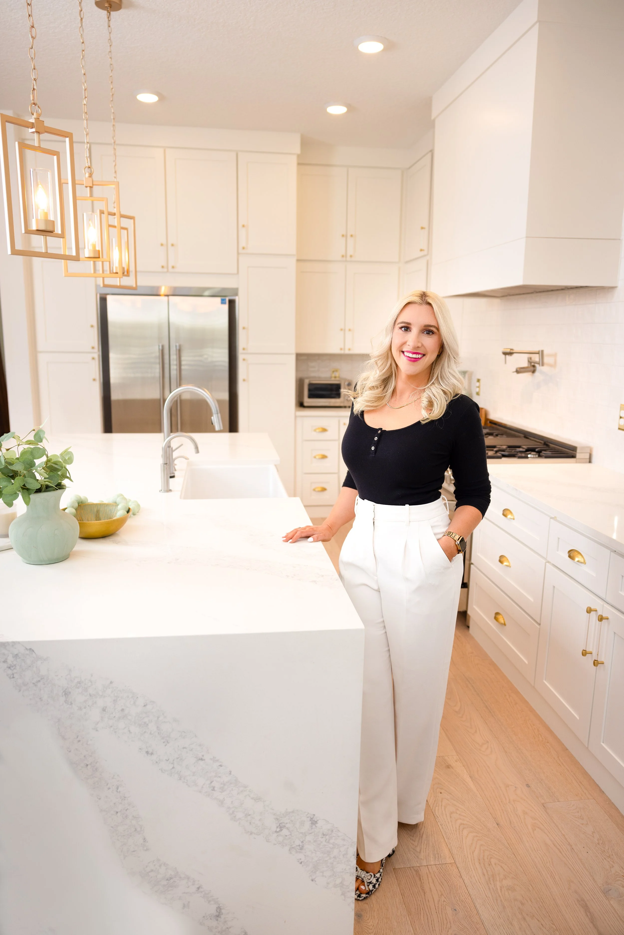 A woman with blonde hair, wearing a black top and white pants, standing in a bright, modern kitchen with white cabinets and gold hardware, smiling at the camera.