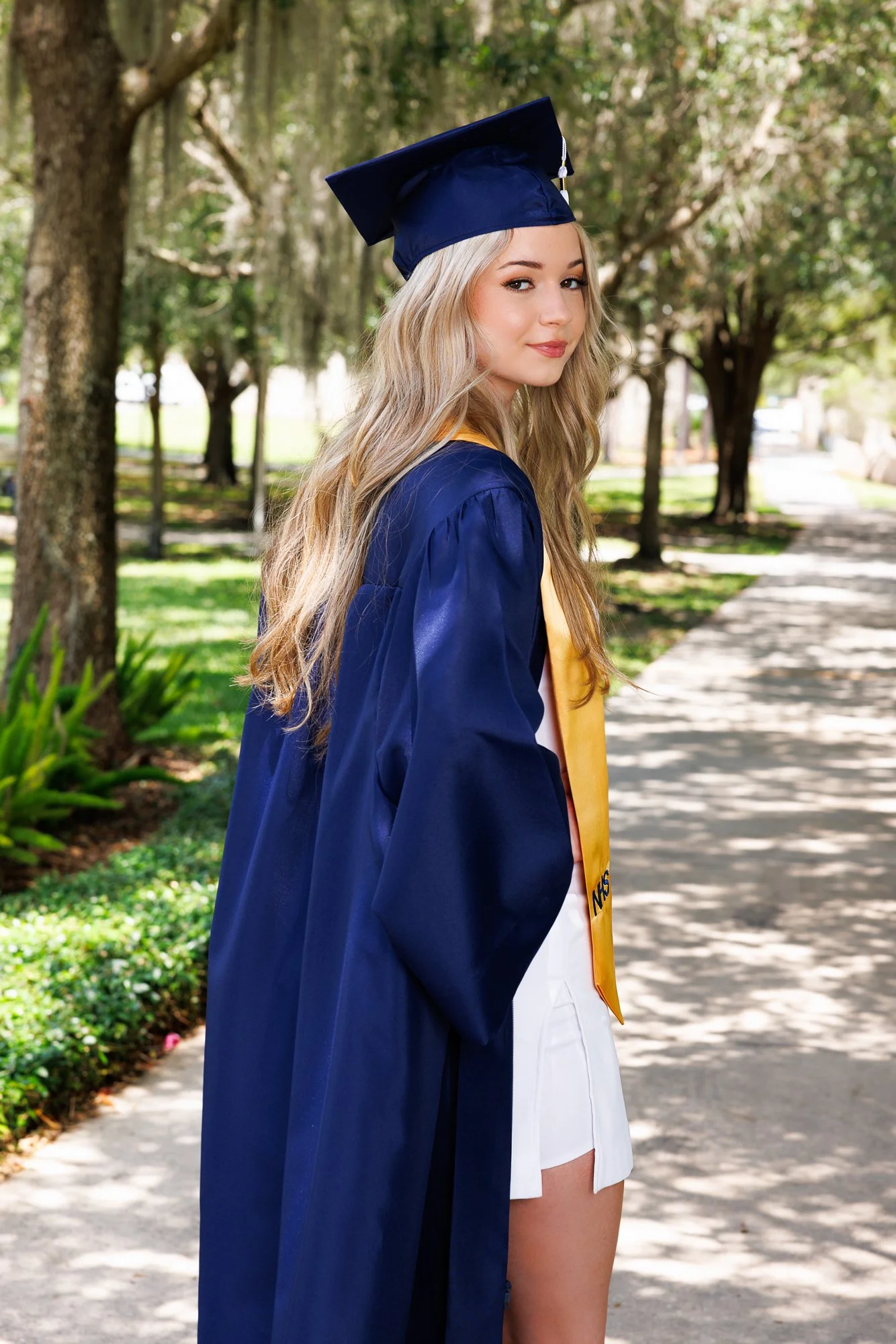 A young woman in a blue graduation gown and cap with a gold stole, standing outdoors on a tree-lined sidewalk during daytime.