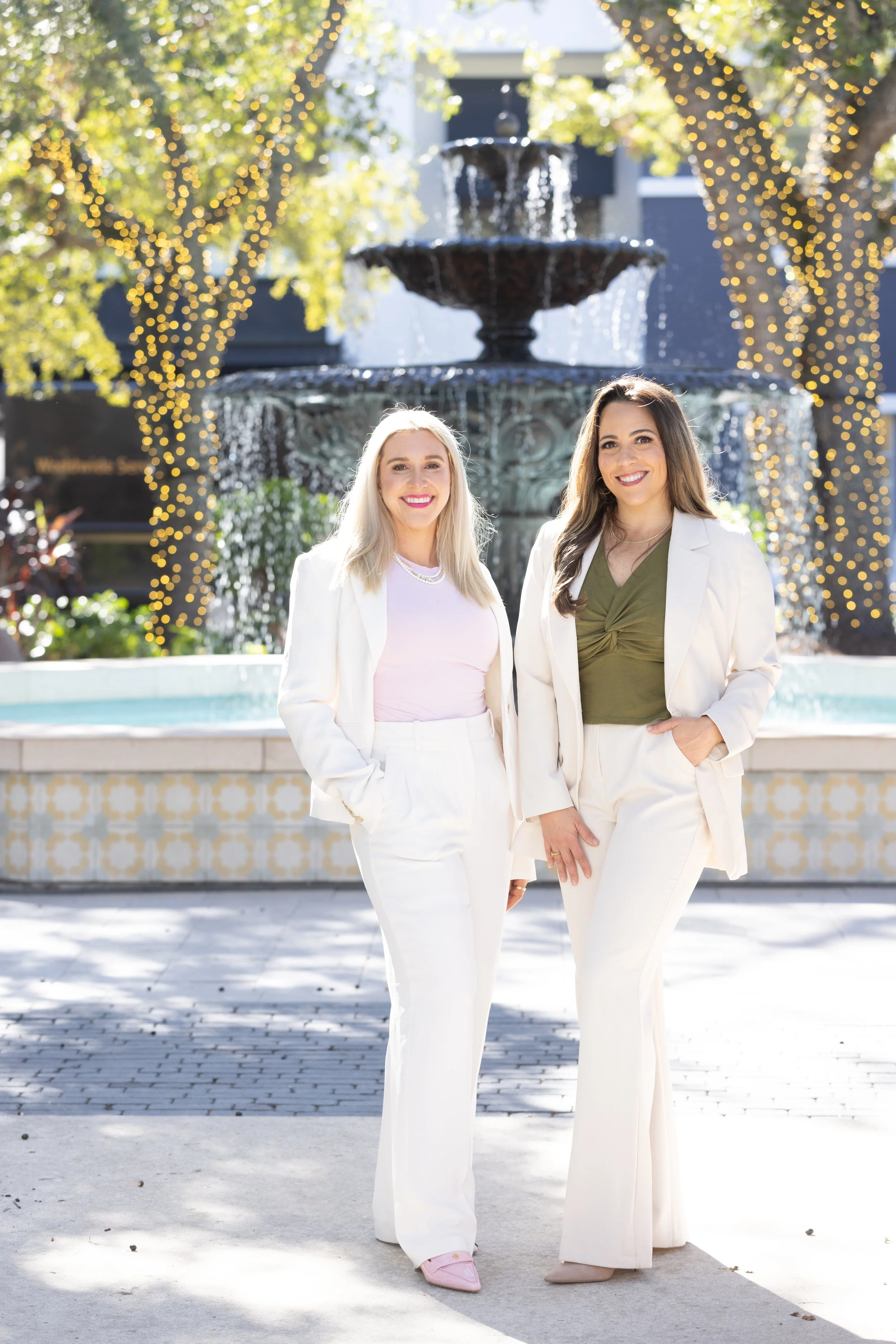Two women standing in front of a fountain with string lights in a park or plaza, dressed in white suits, smiling at the camera.