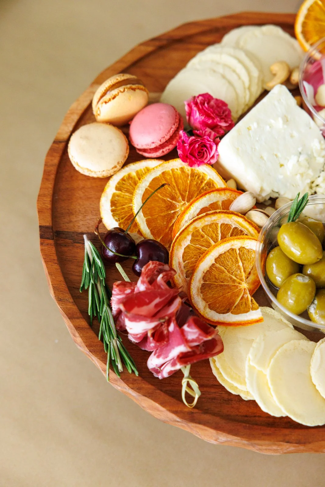 Wooden charcuterie board with cheese, dry cured meats, orange slices, cherries, green olives, pink and white macarons, pink flowers, and a sprig of rosemary.