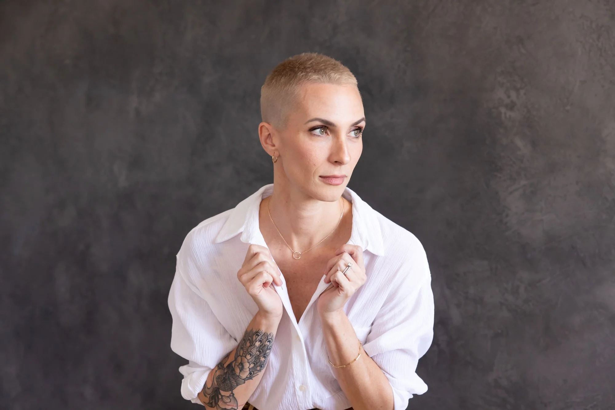 A woman with a short blonde haircut wearing a white shirt, jewelry, and earrings, holding her collar with both hands, looking to her left against a dark gray background.