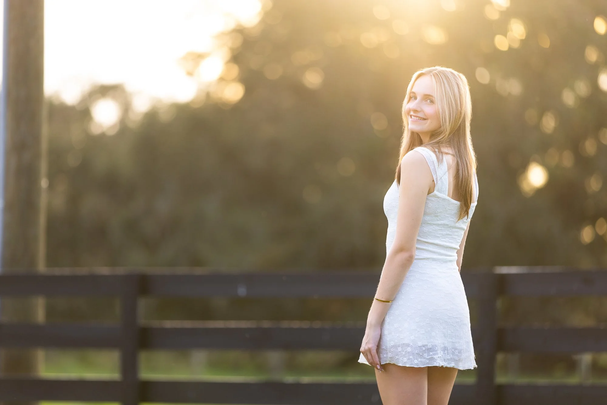 Young woman in a white dress smiling outdoors in the late afternoon or early evening.