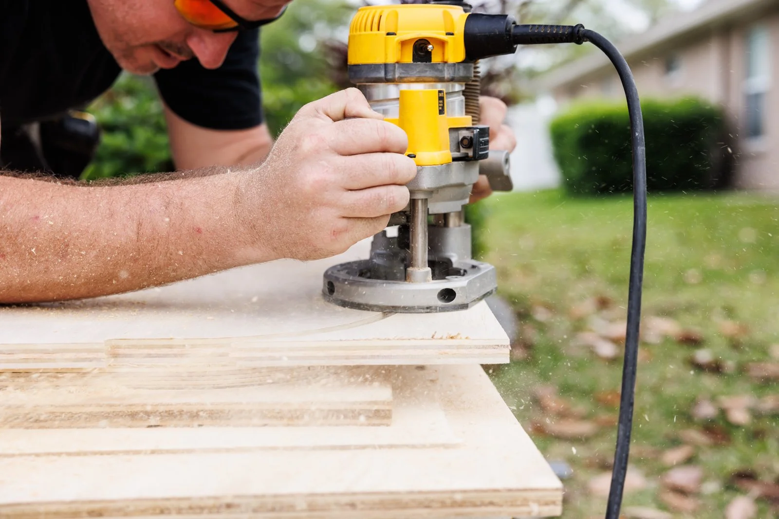 A man using a yellow and gray router to carve or cut a piece of wood outdoors, with wood scraps flying around.