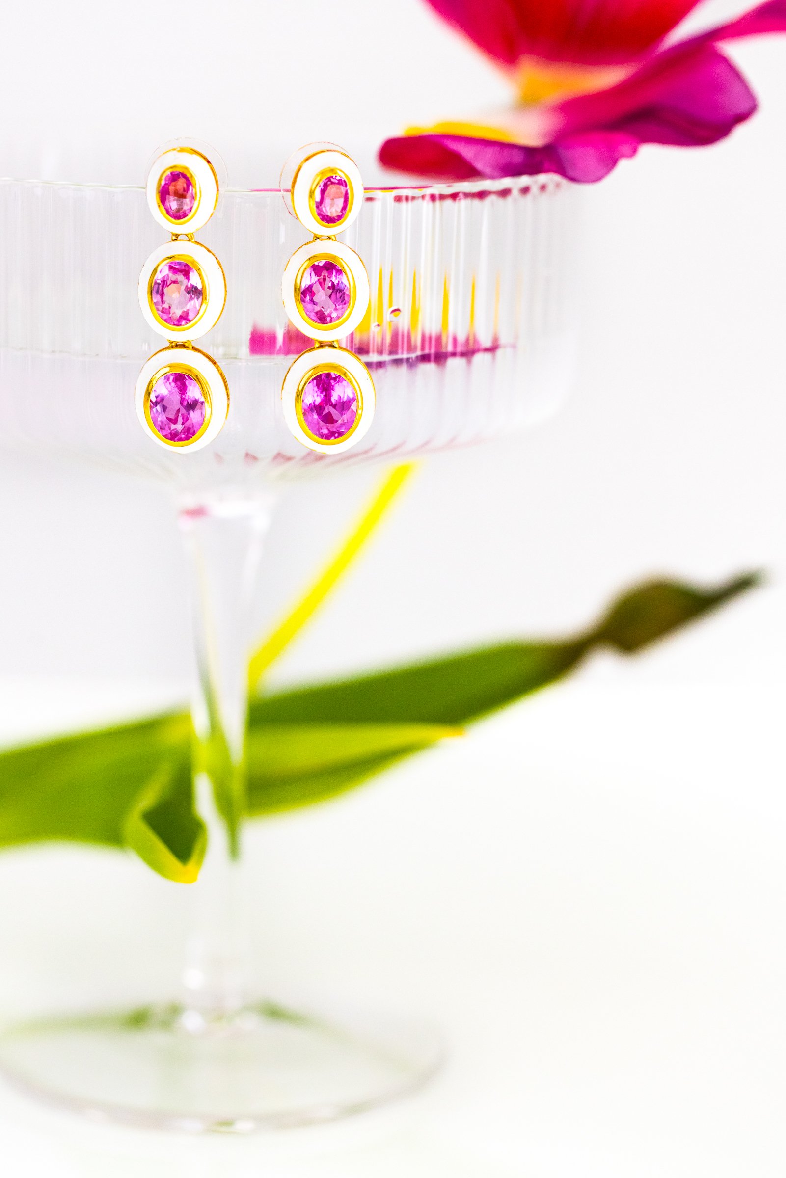 Pink gemstone drop earrings hanging on a clear glass dish with a pink flower and green leaves.
