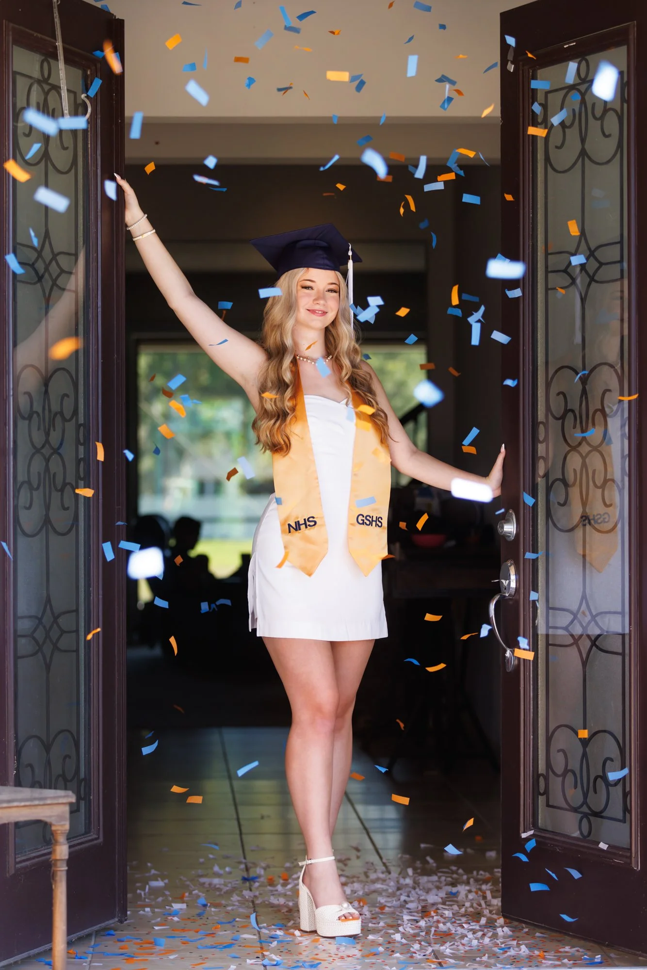 A young woman in a graduation cap and gown, with a yellow stole, celebrating in a doorway as colorful confetti falls around her.