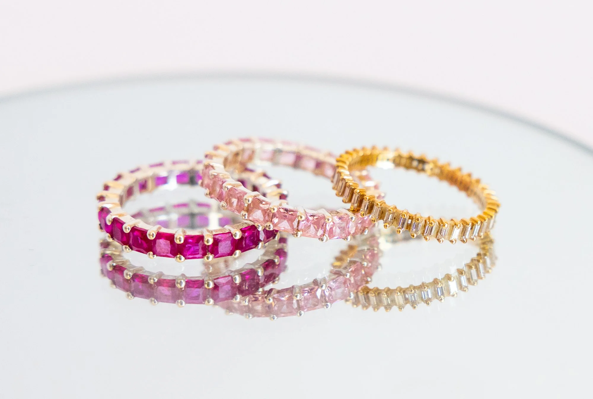 Three rings with pink and red gemstones and gold bands on a reflective surface.