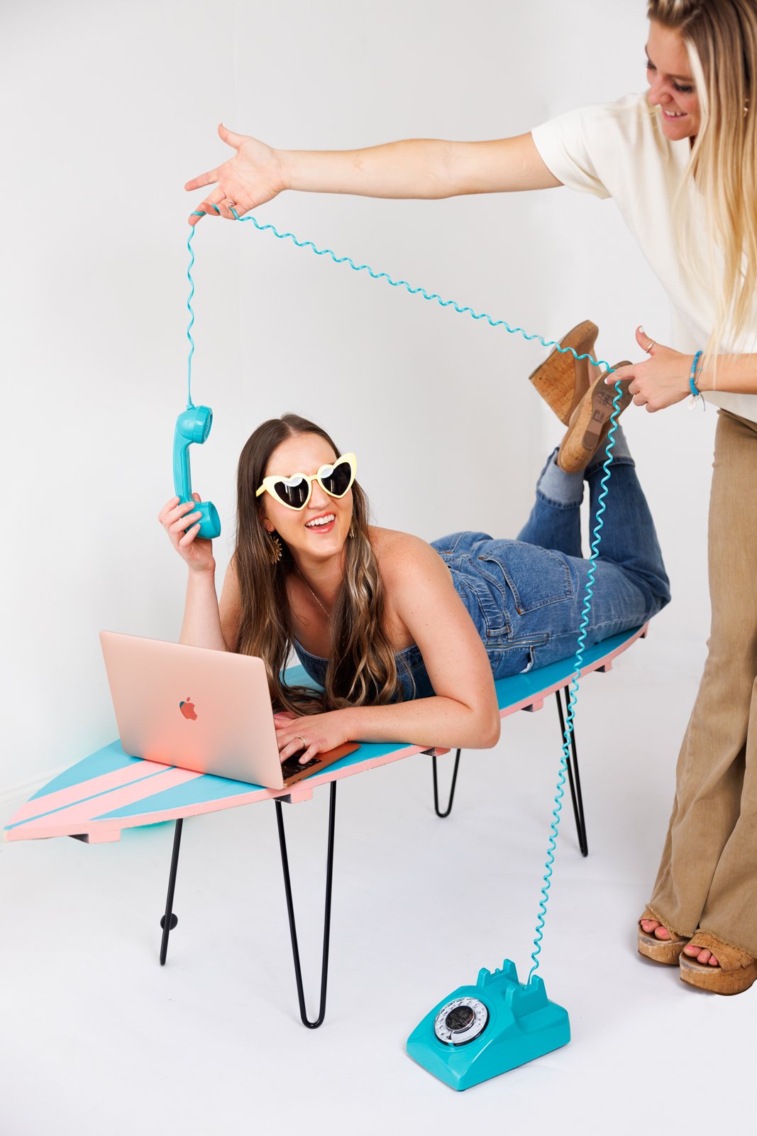 A woman lying on a surfboard with a laptop, wearing heart-shaped sunglasses, holding a blue retro rotary phone, and smiling while another person drags a blue cord from the phone to her.