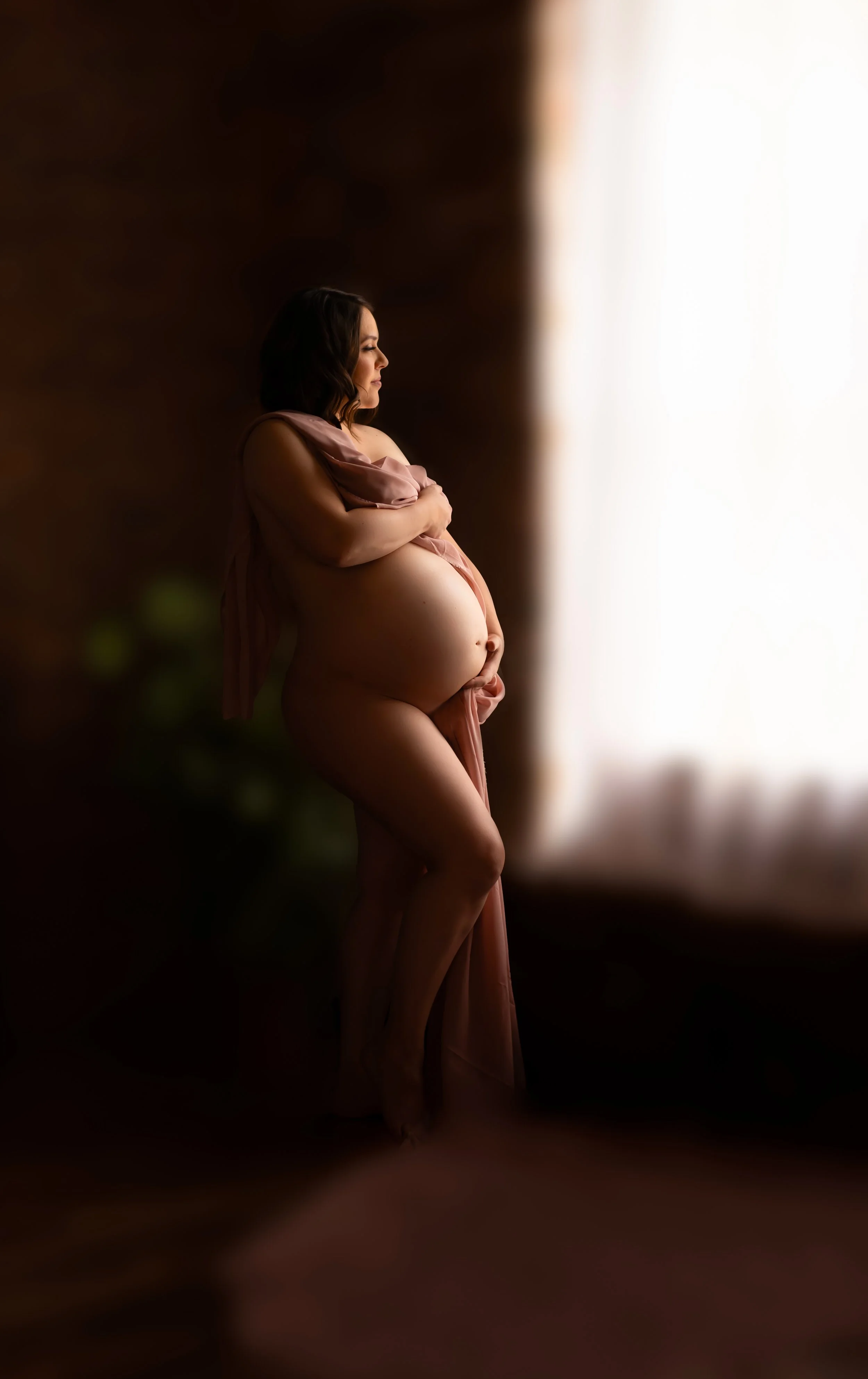 Pregnant woman standing by a window with soft natural light, holding her belly in a gentle pose.