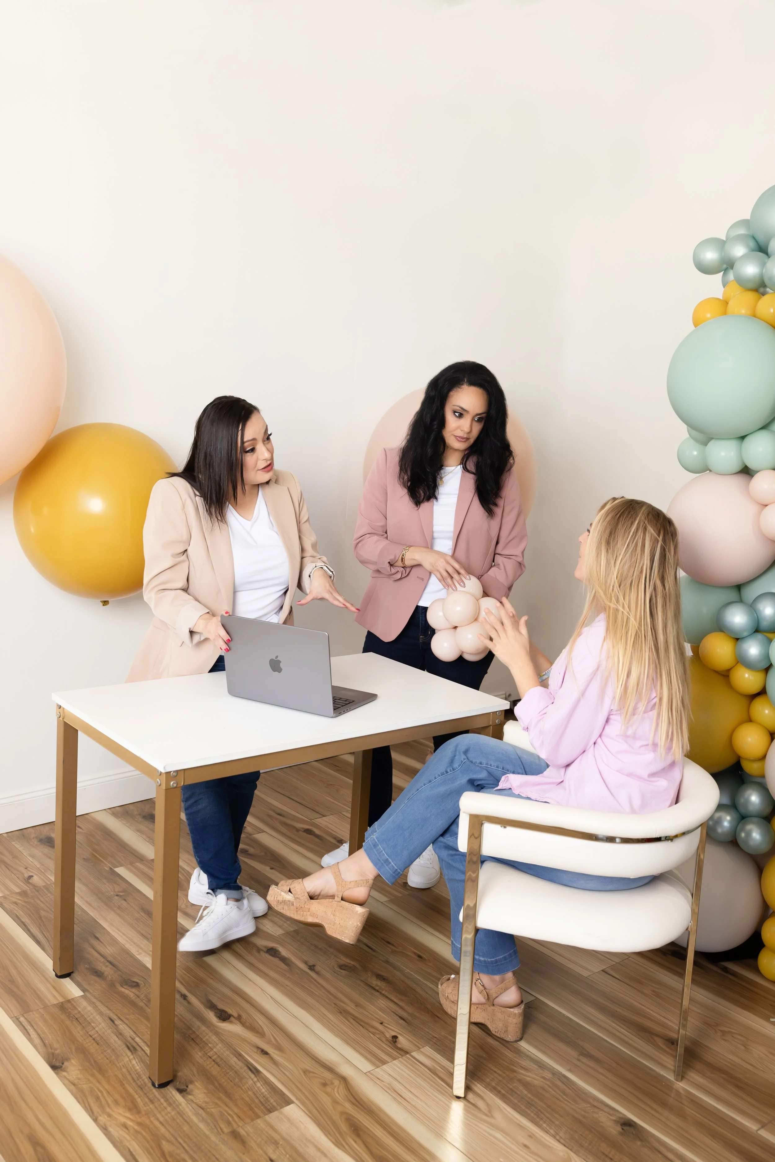 Three women are in a room decorated with pastel-colored balloons. One woman is sitting in a white chair, showing a balloon arrangement to two standing women. One standing woman is pointing, and the other is holding balloons. There is a laptop on a wh