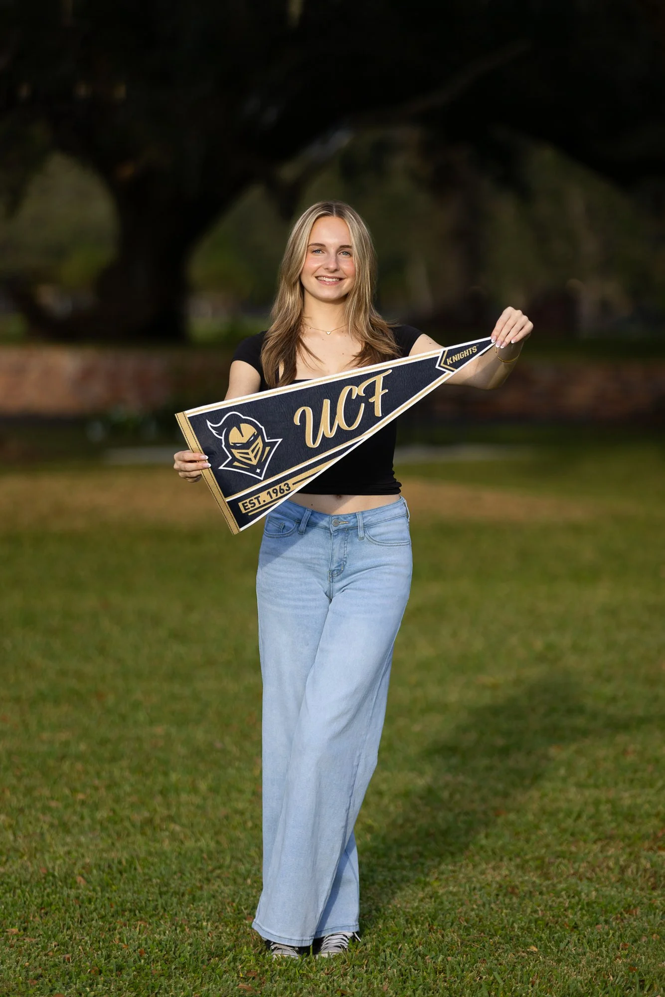 A young woman with long blonde hair, wearing a black crop top and light blue jeans, standing outdoors on grass in front of a large tree, holding a university flag with the letters 'UCF' and a Knight logo.