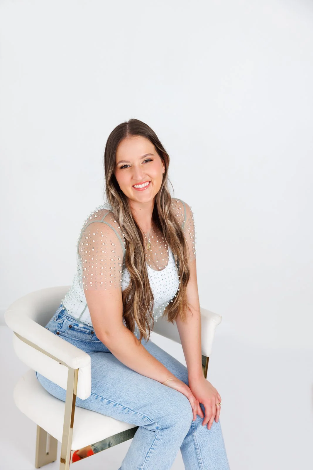 A young woman with long brown hair, smiling, sitting on a white chair against a plain white background, wearing a sheer top with pearl embellishments and light blue jeans.