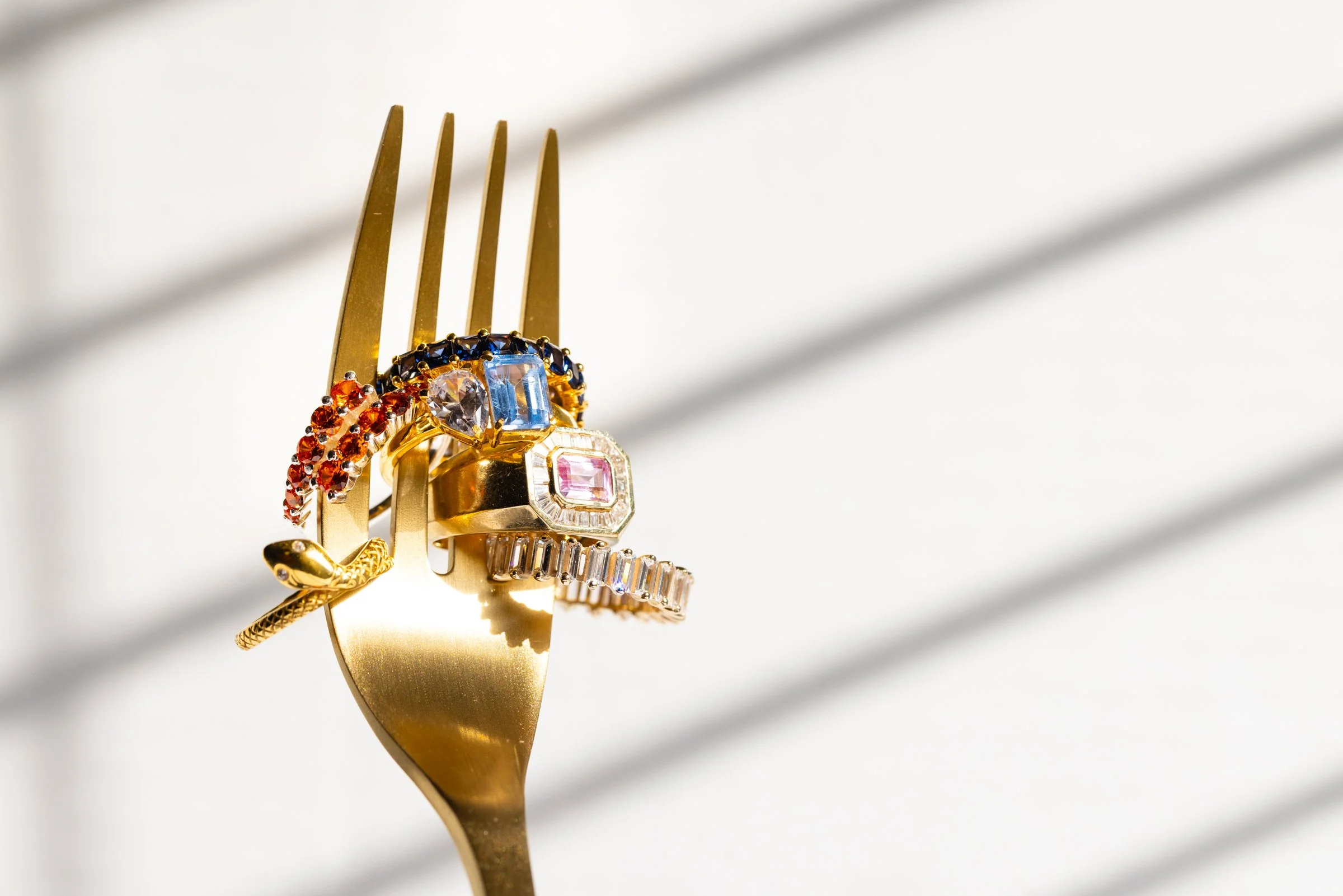 Gold fork with multiple colorful gemstone rings hanging on its prongs, casting shadows on a white surface.
