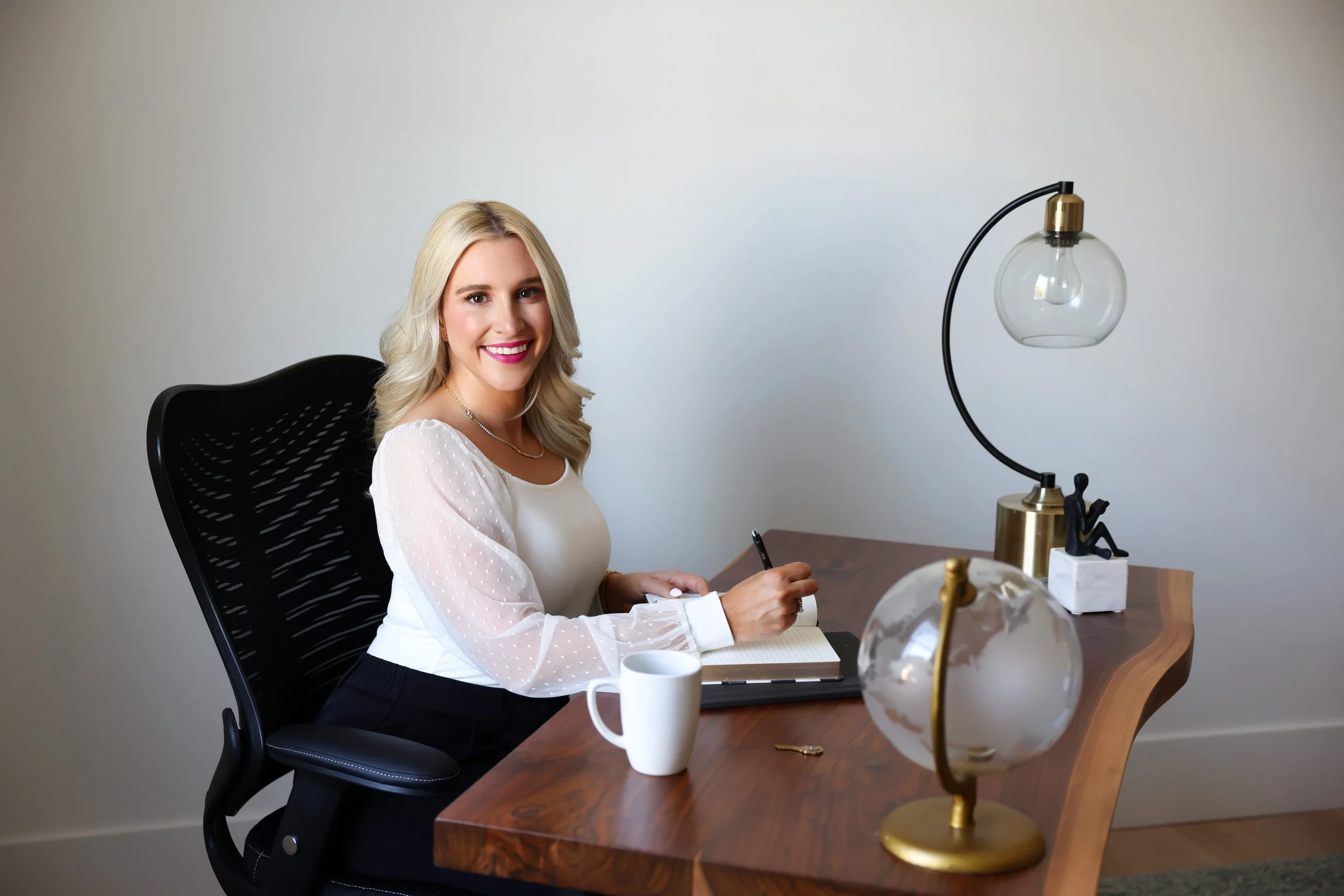 A woman with blonde hair and a white blouse sitting at a wooden desk, smiling, with a white mug, a globe, and a lamp on the desk.