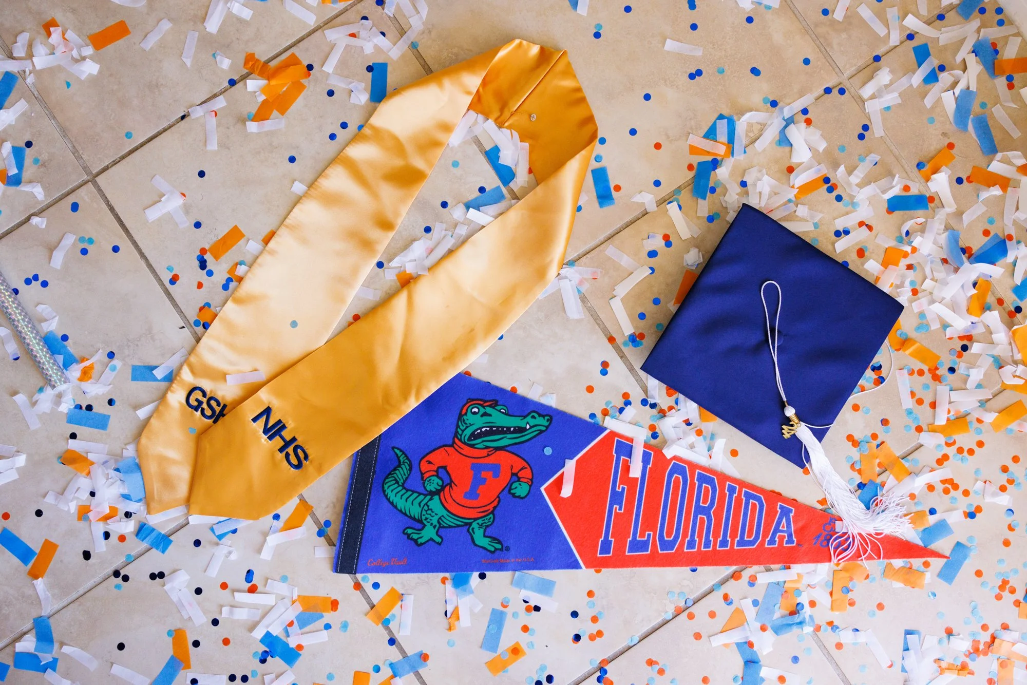 A celebration scene with confetti, a Florida Gators banner, a navy blue graduate cap with tassel, and a yellow sash with blue text on a tiled floor.