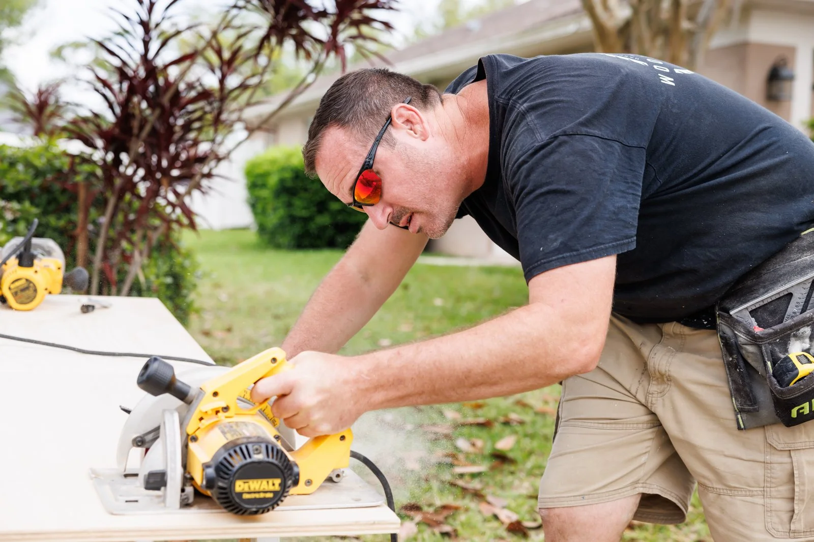 Man cutting wood with a yellow DeWalt miter saw outside in a yard, wearing sunglasses, black t-shirt, and khaki shorts.