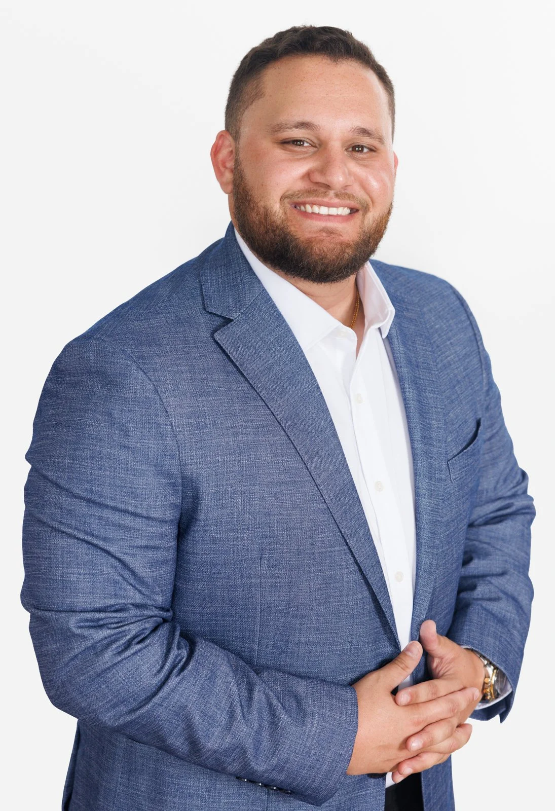 A man with a beard smiling, wearing a blue blazer and white shirt, standing against a white background.