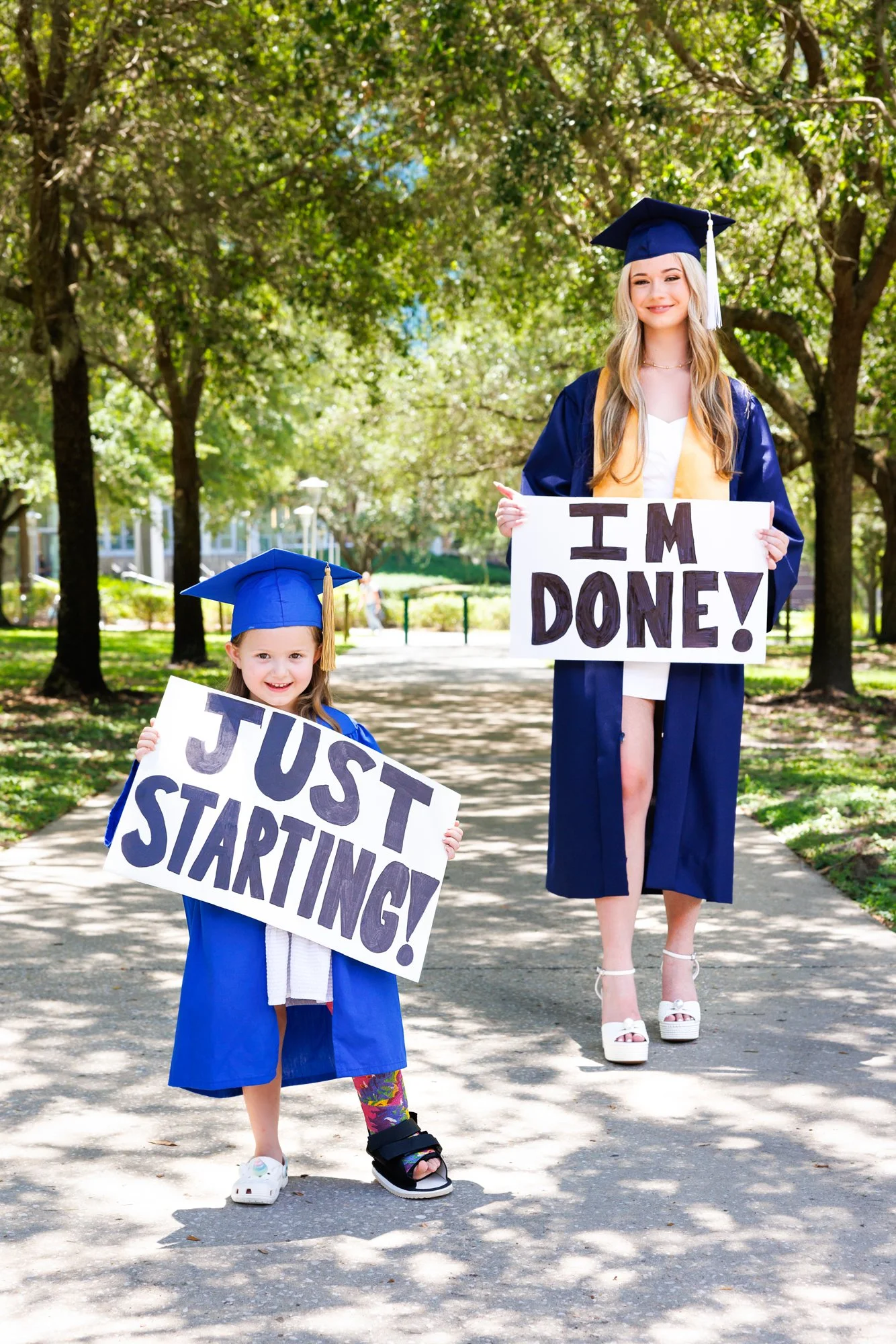 A young girl and a woman in graduation caps and gowns holding signs. The girl’s sign says 'JUST STARTING!' and the woman’s sign says 'I'M DONE!'. They are standing on a tree-lined path outside.