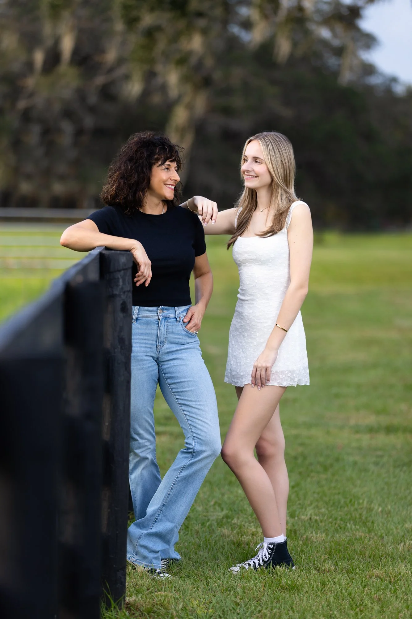 Two women, one in a black shirt and jeans, and the other in a white dress with black sneakers, are standing outdoors on a grassy field, smiling and talking, with trees in the background.